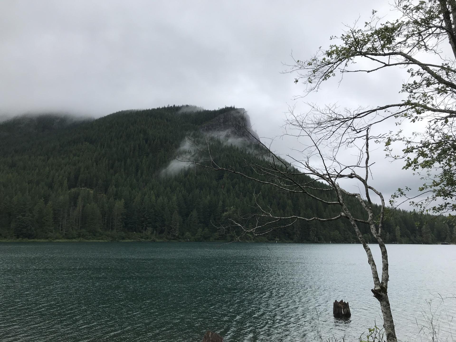 A dark lake in front of a cloud-covered, forested mountain on an overcast day.