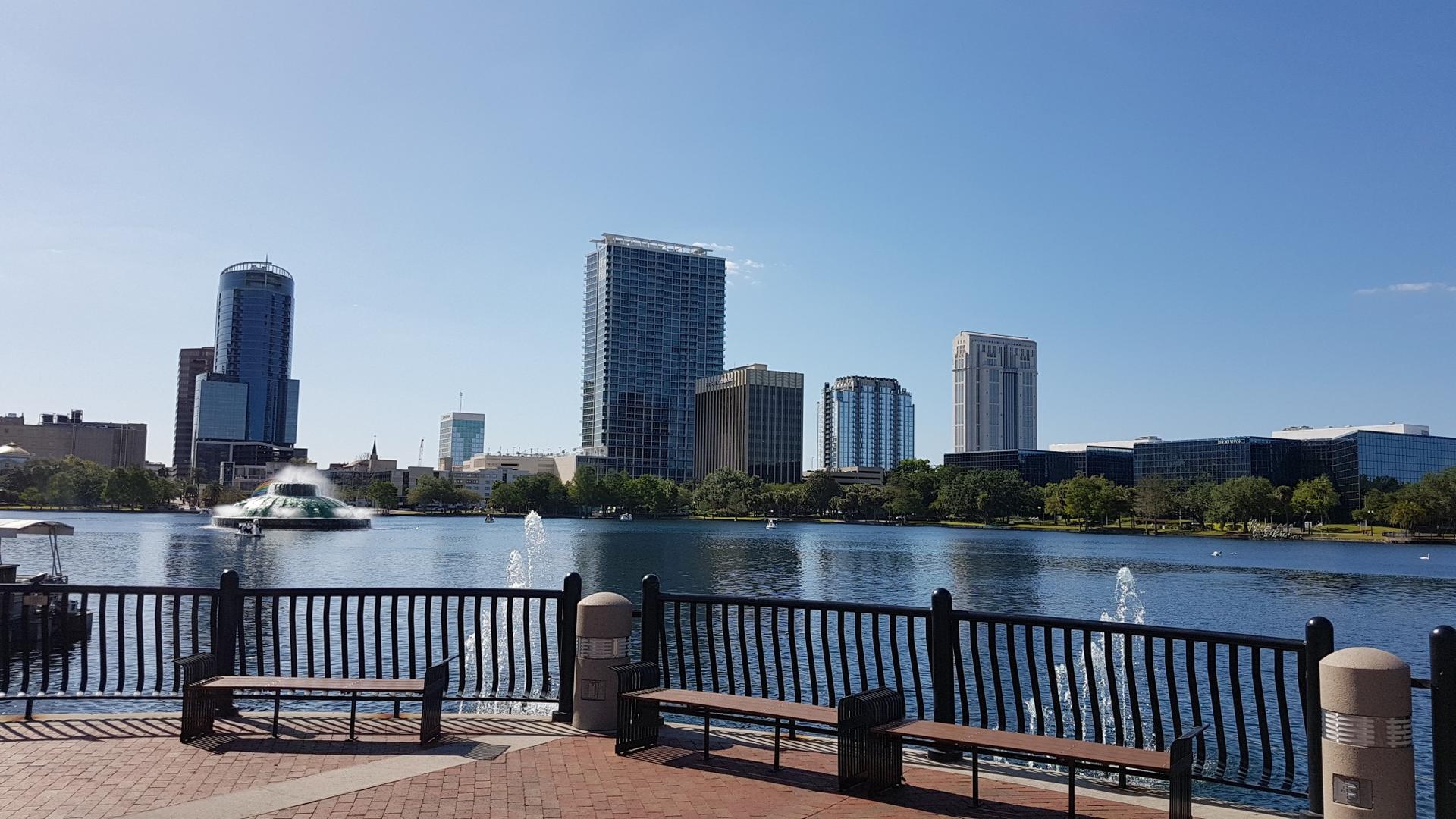 City skyline across a lake with fountains, seen from a waterfront promenade.