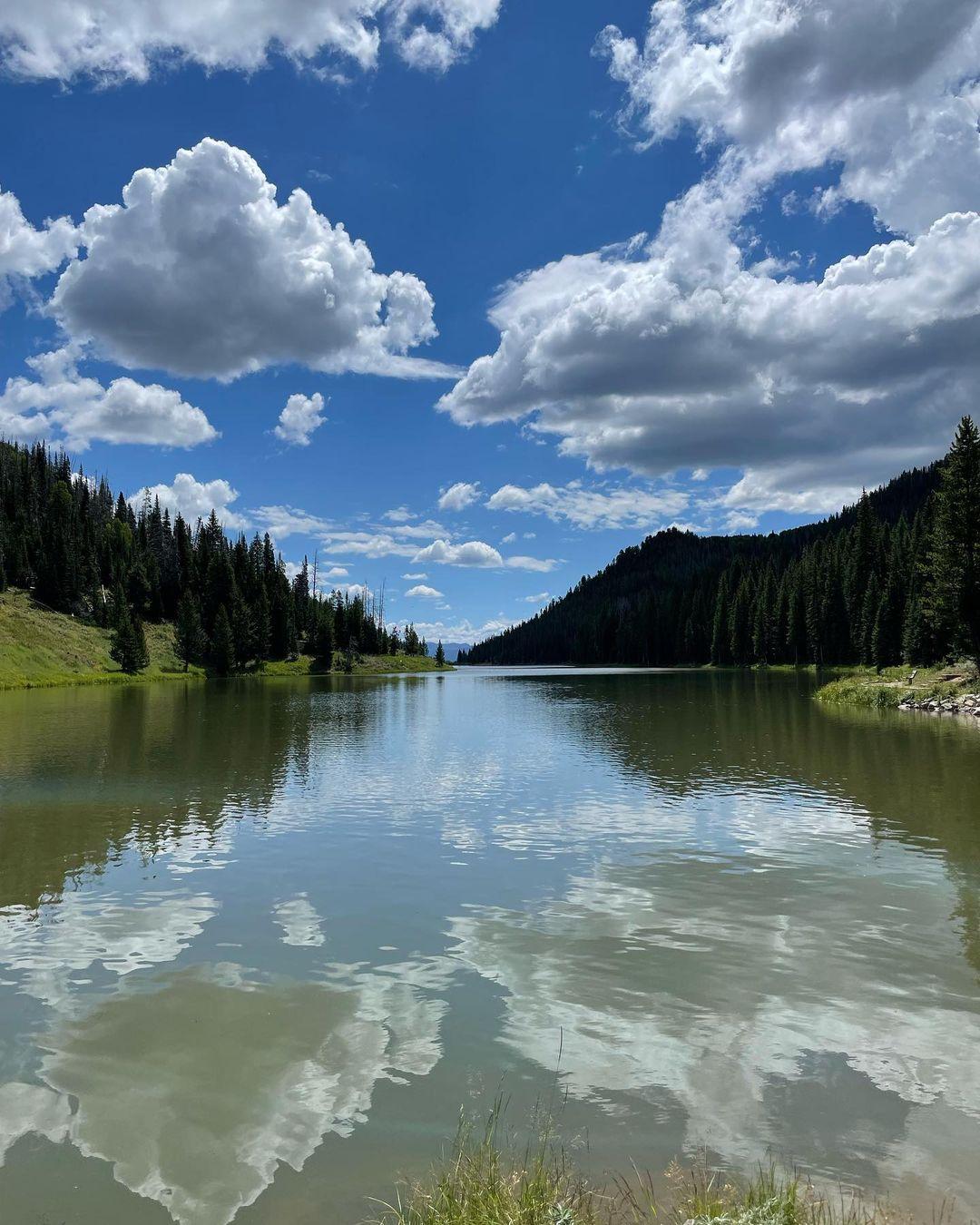 Calm lake reflecting blue sky and white clouds, surrounded by pine hills.
