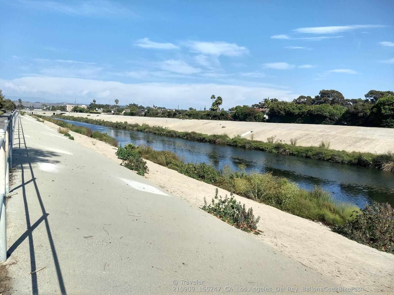 Sunny concrete channel with flowing water and green vegetation under blue sky.