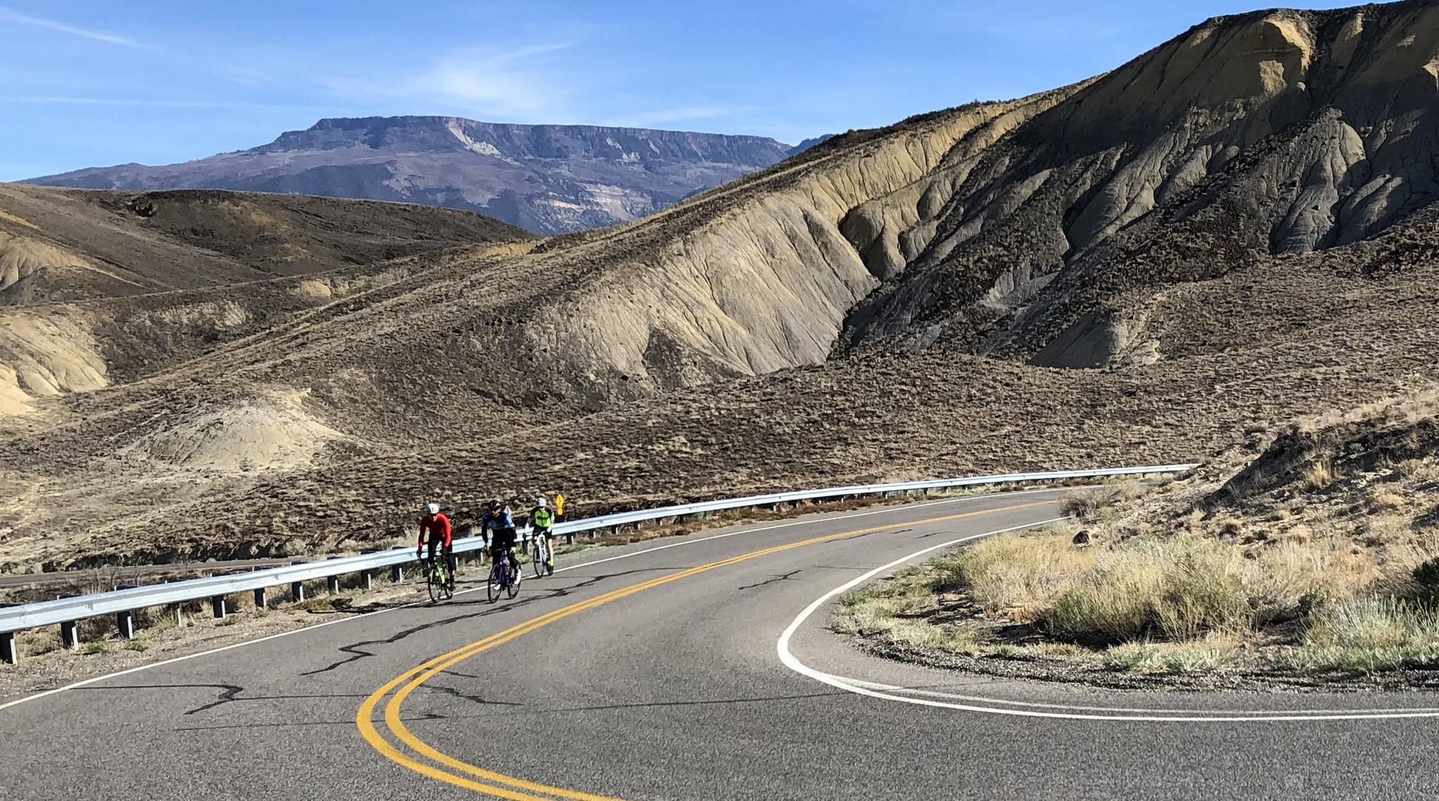 Three cyclists ride on a winding road through arid, rolling hills. - Fruit and Wine Byway Loop
