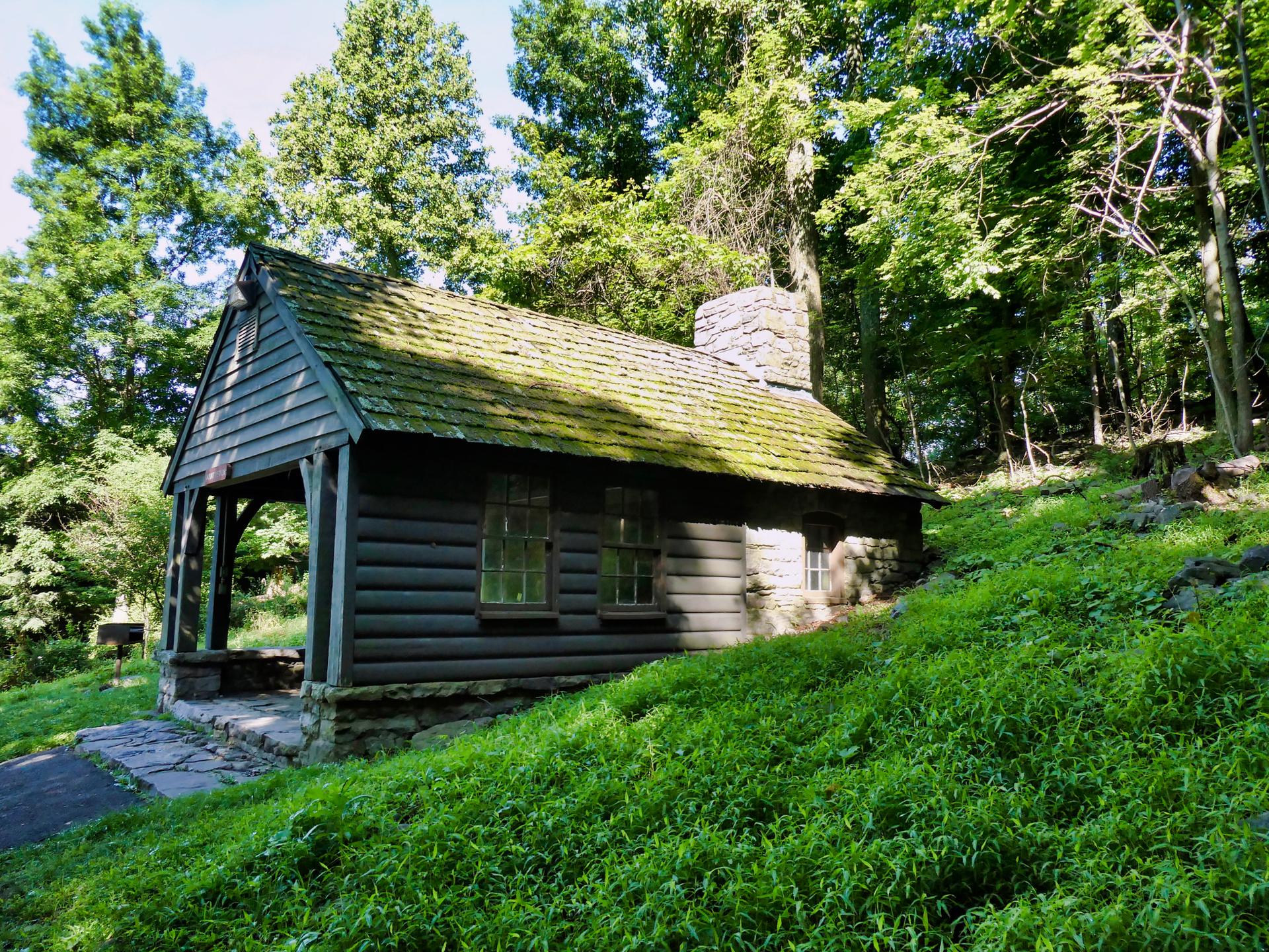 Rustic wooden cabin with stone chimney nestled on a sunny, green hillside.