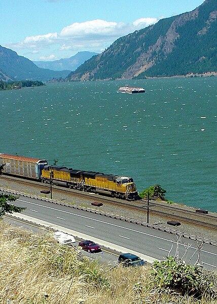 A yellow train on tracks beside a highway, a wide river, and green mountains.