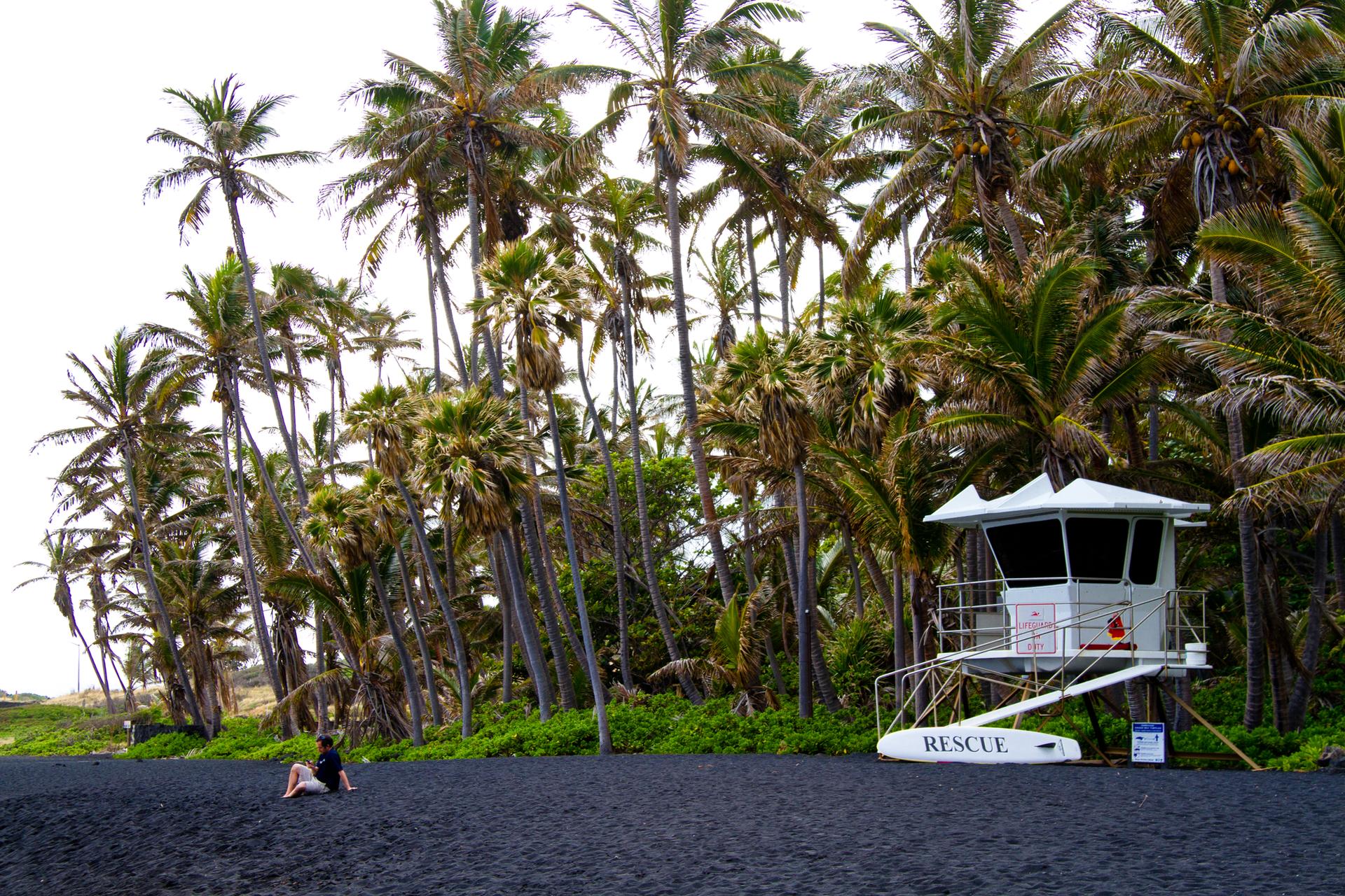 Person sits on black sand beach near a white lifeguard tower and palm trees.