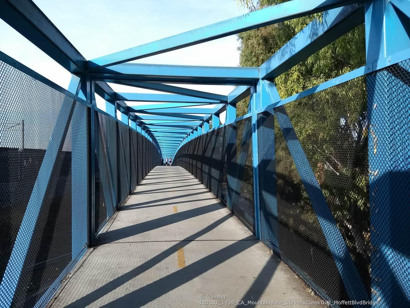 A long blue pedestrian bridge with strong shadows from bright sunlight.