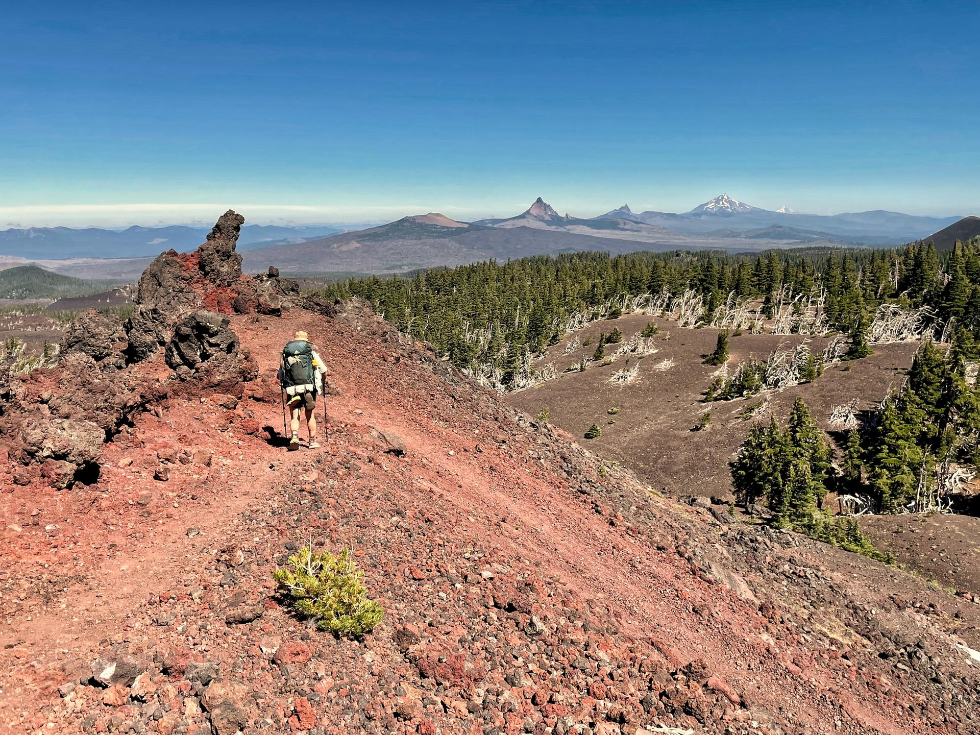 A hiker with a backpack walks along a red volcanic ridge on a sunny day.