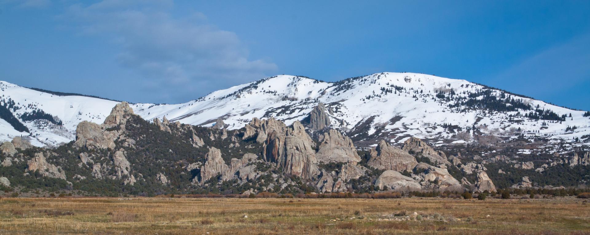 Panoramic view of snow-capped mountains, rocky formations, and a dry field.