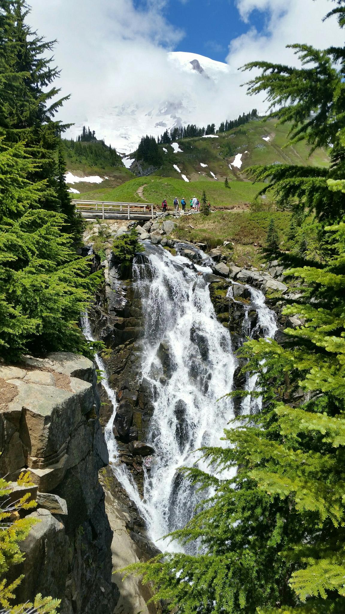A multi-tiered waterfall cascades down rocks, with a snowy mountain in the background.