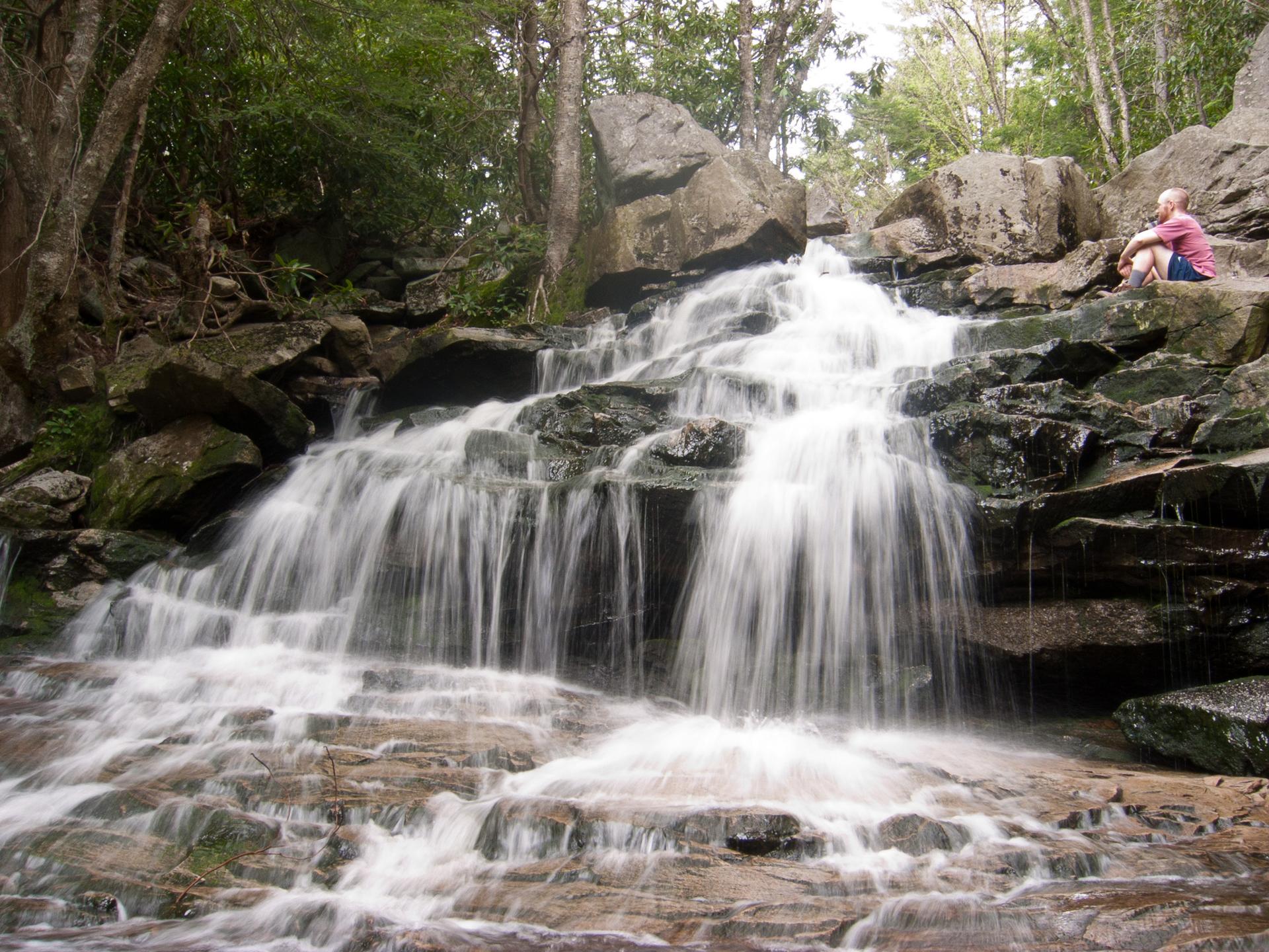 A person sits beside a multi-tiered waterfall in a rocky, forested landscape.