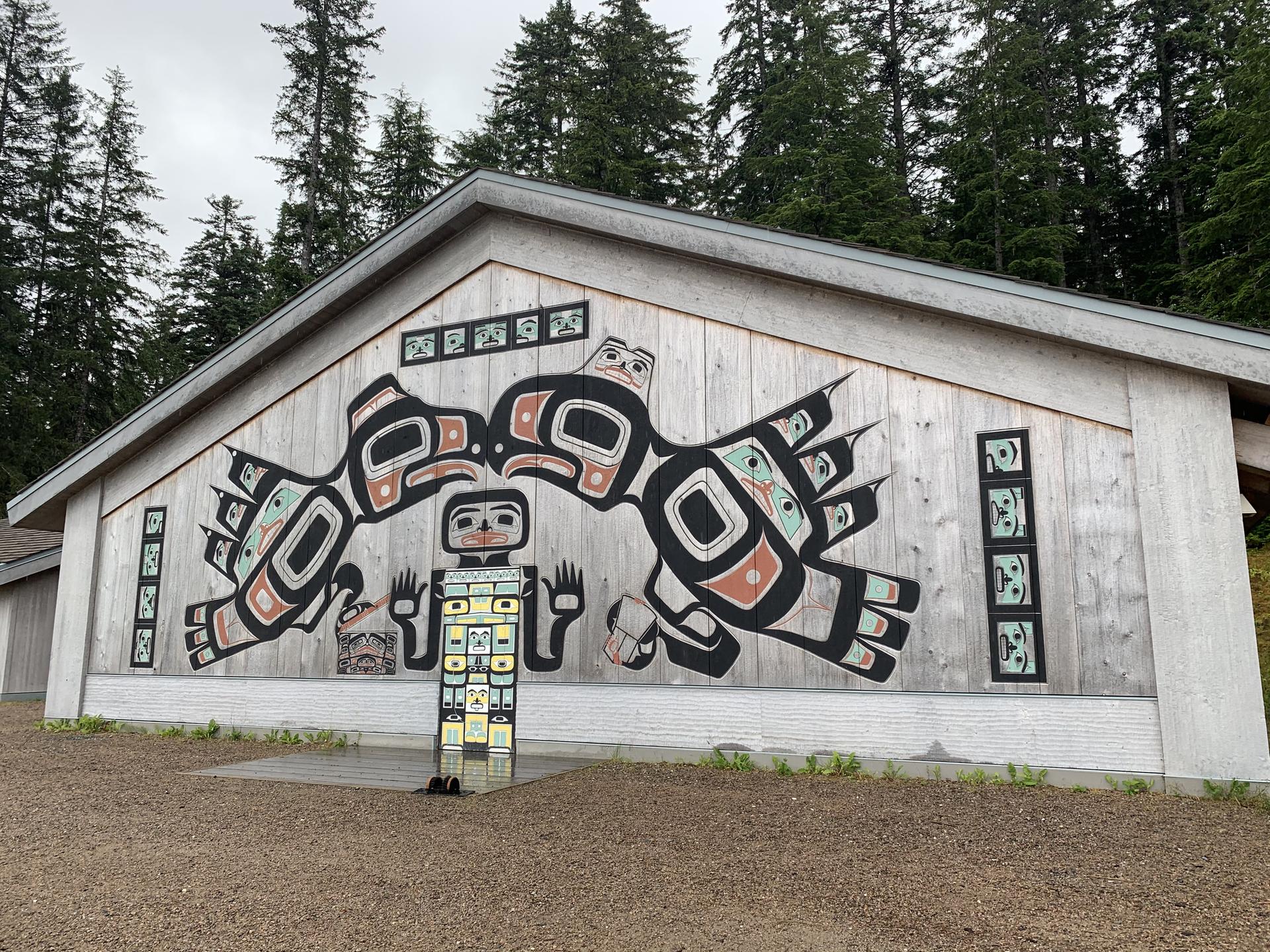 A building with a large, colorful mural and a totem pole in front of a forest.