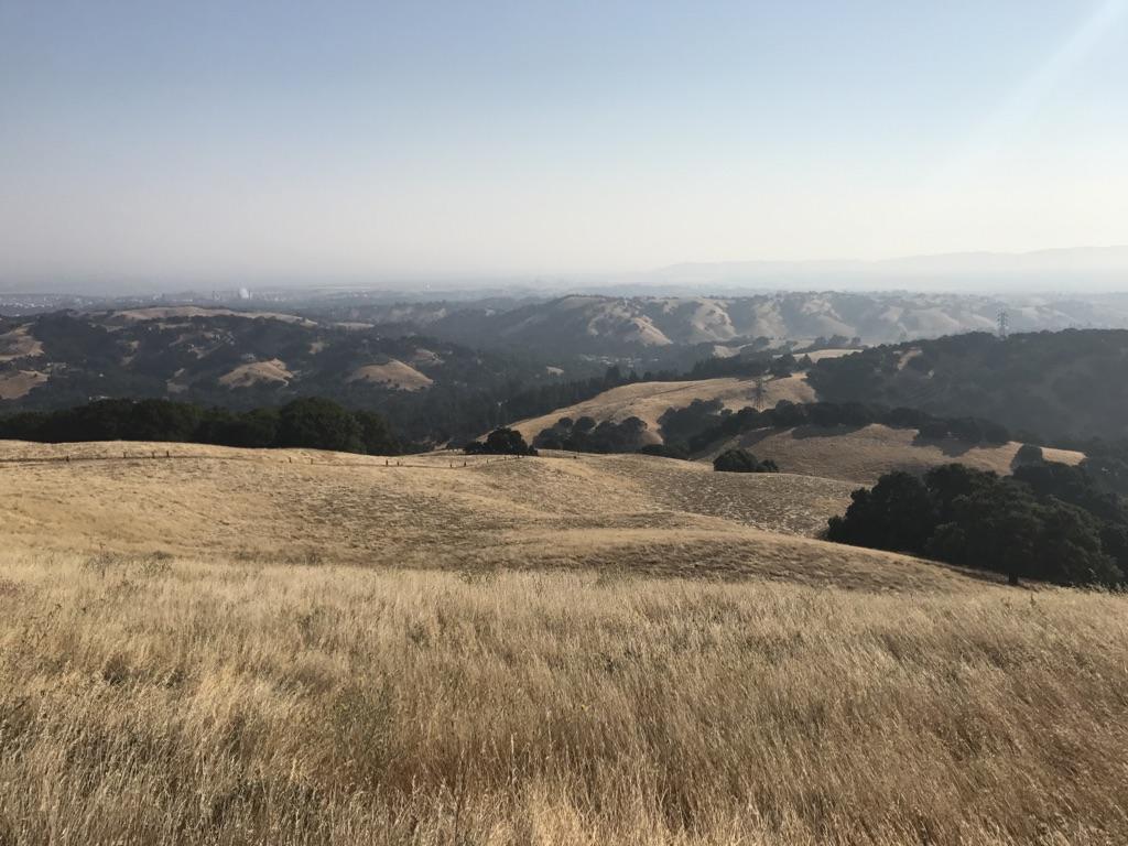 Expansive view of golden dry hills and distant hazy mountains under a clear sky.