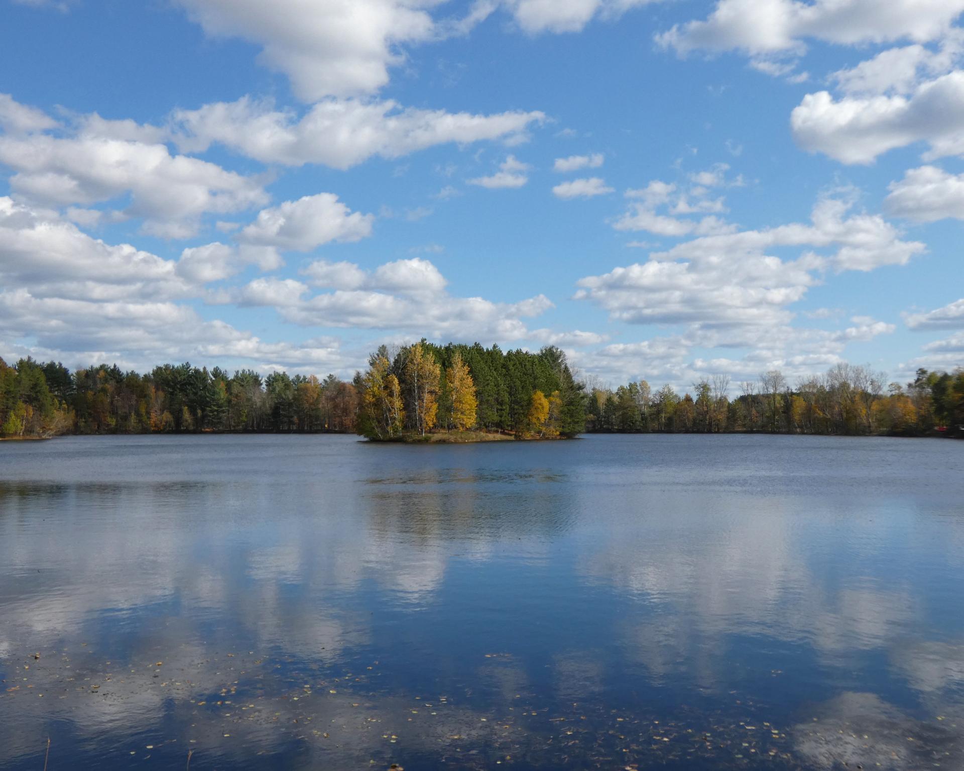 A calm lake reflects a blue sky and white clouds, surrounded by autumn trees.