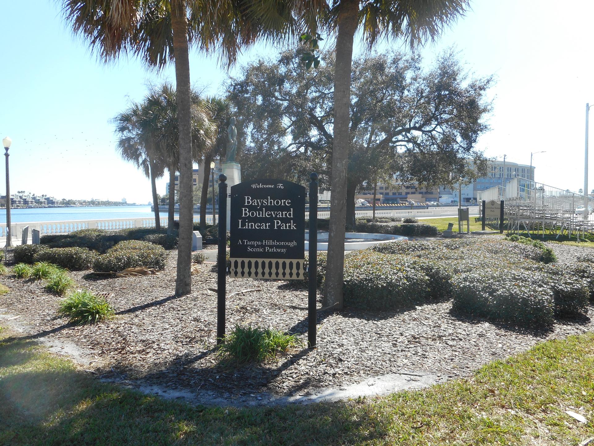 Park sign surrounded by palm trees and bushes with water in the background.