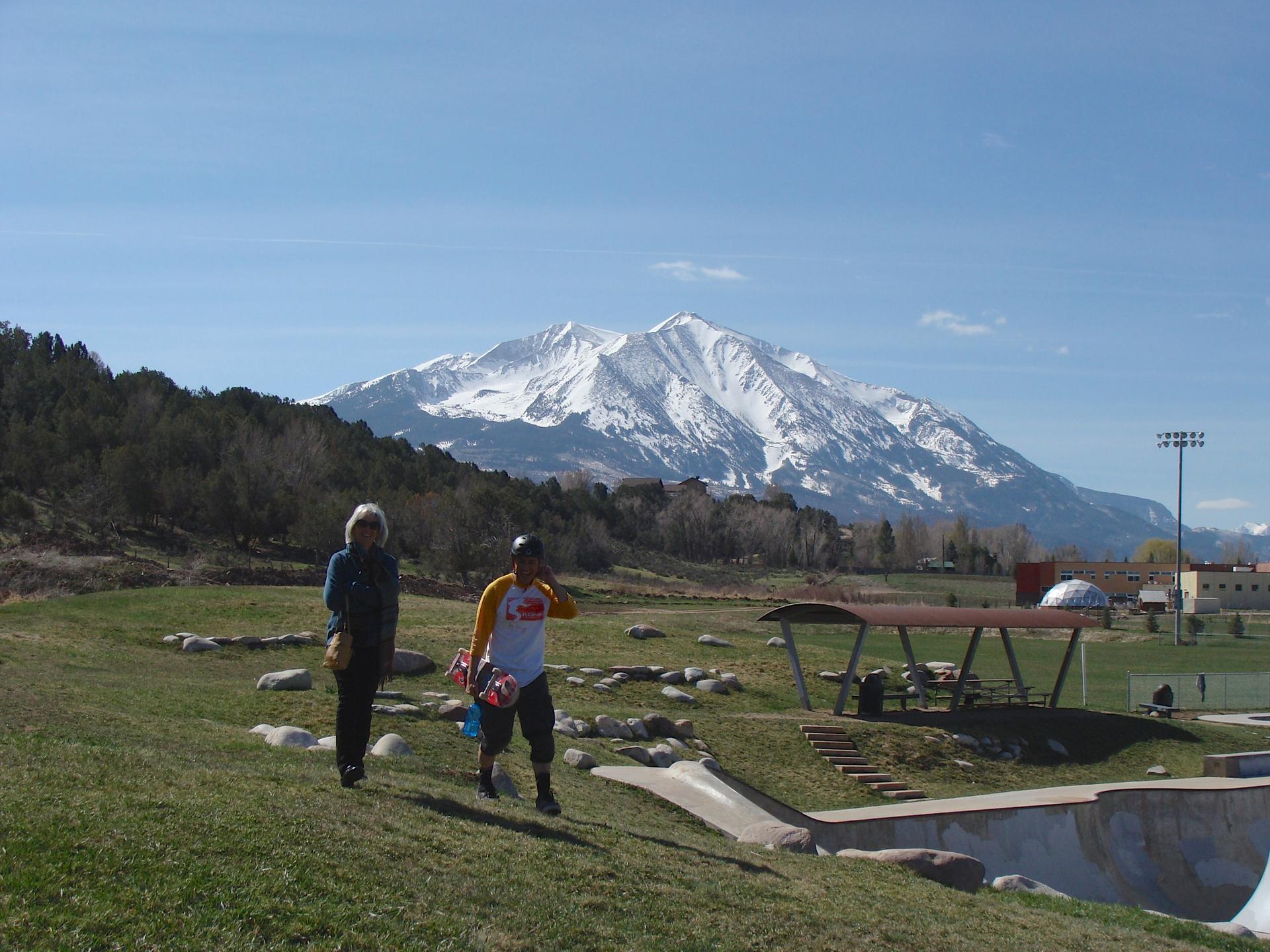 Two people walk on a sunny grassy hill with a snow-capped mountain in the distance.