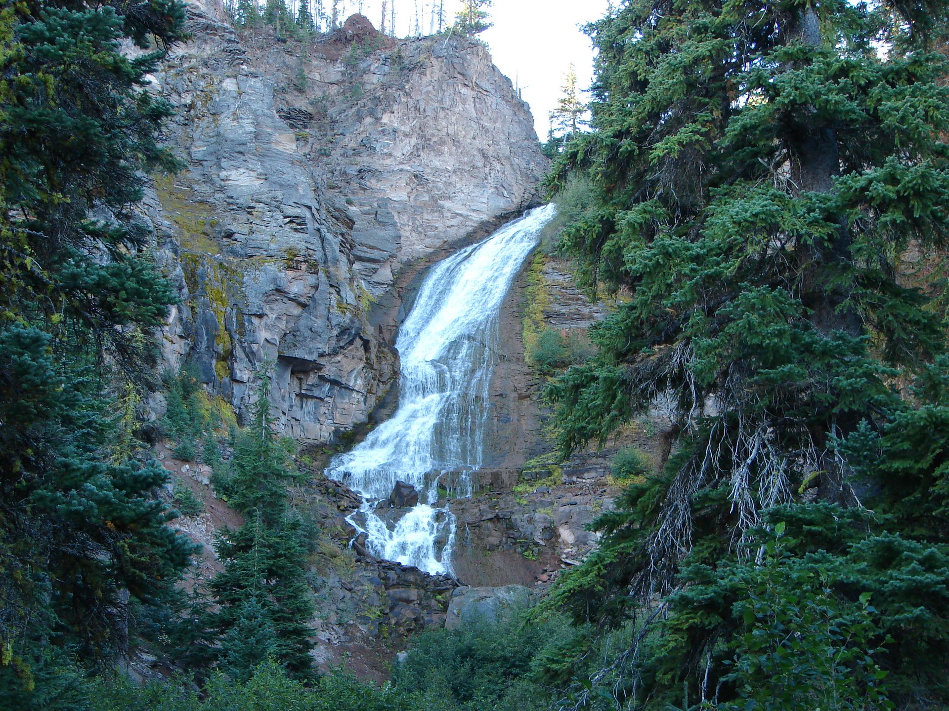 A tall waterfall plunges down a rocky cliff face, framed by green trees.