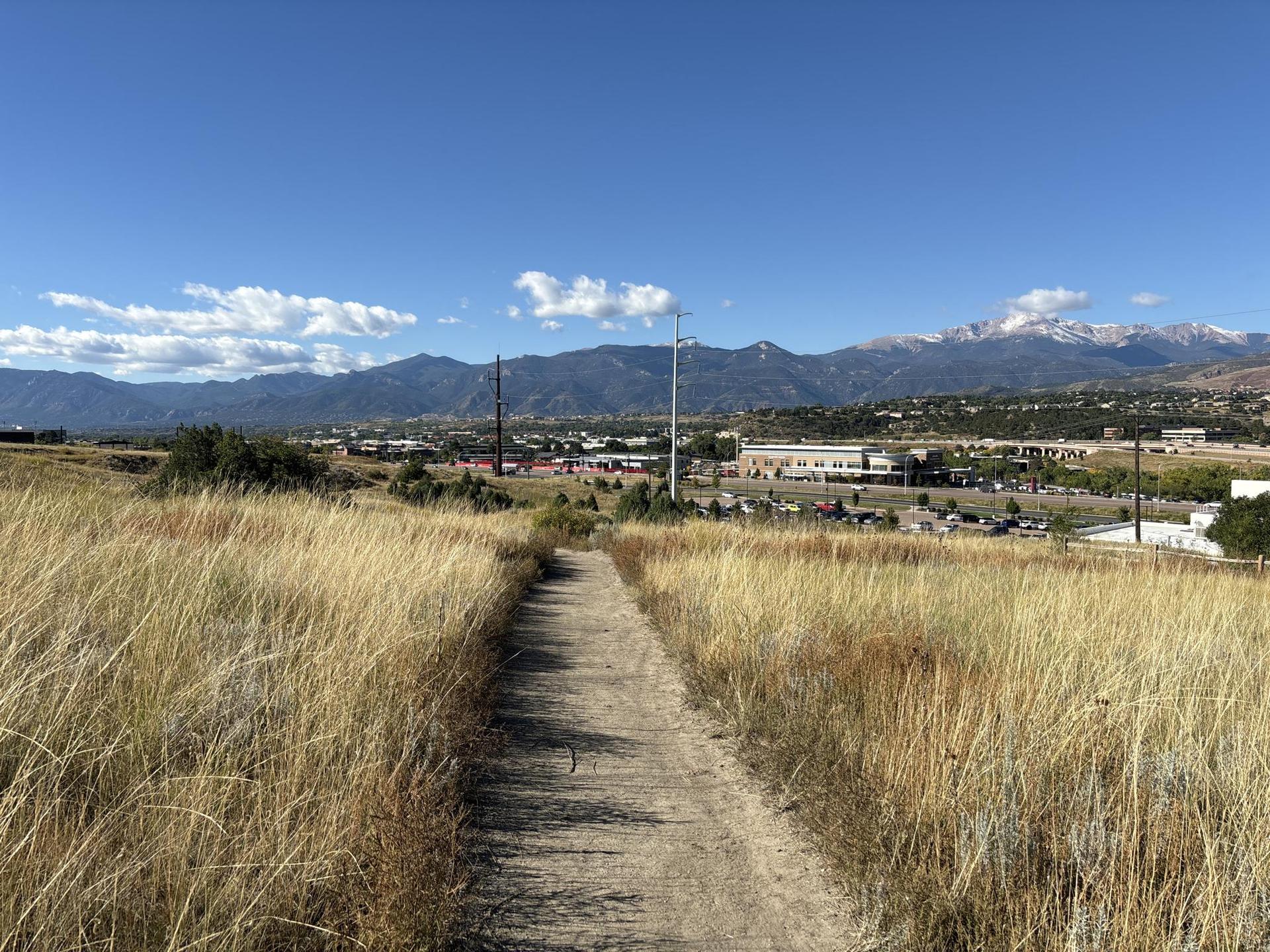 A dirt path through golden grass leads to distant mountains and buildings.