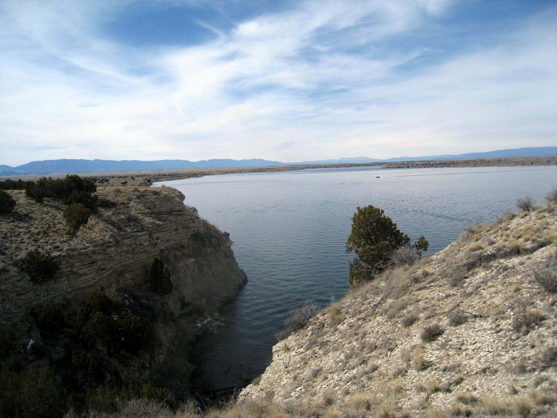 Expansive lake surrounded by arid, rocky cliffs and distant mountains under a bright sky.