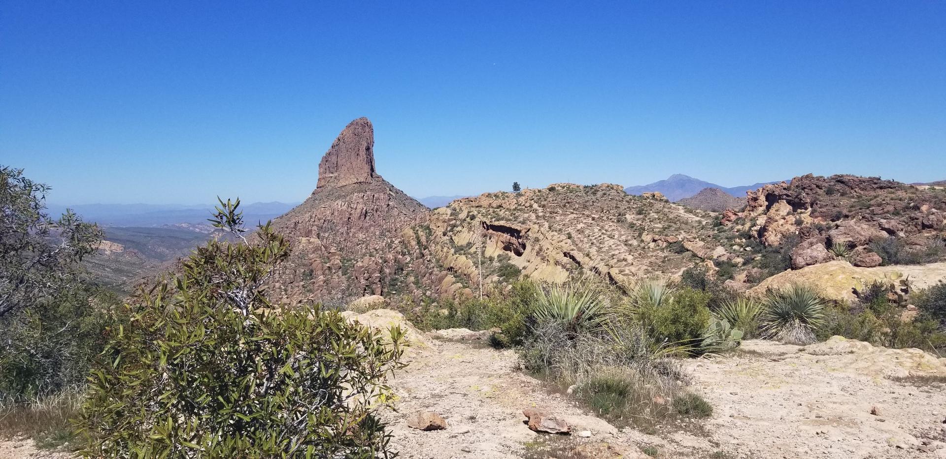 Sunny desert landscape with a prominent rock spire and distant hiker.