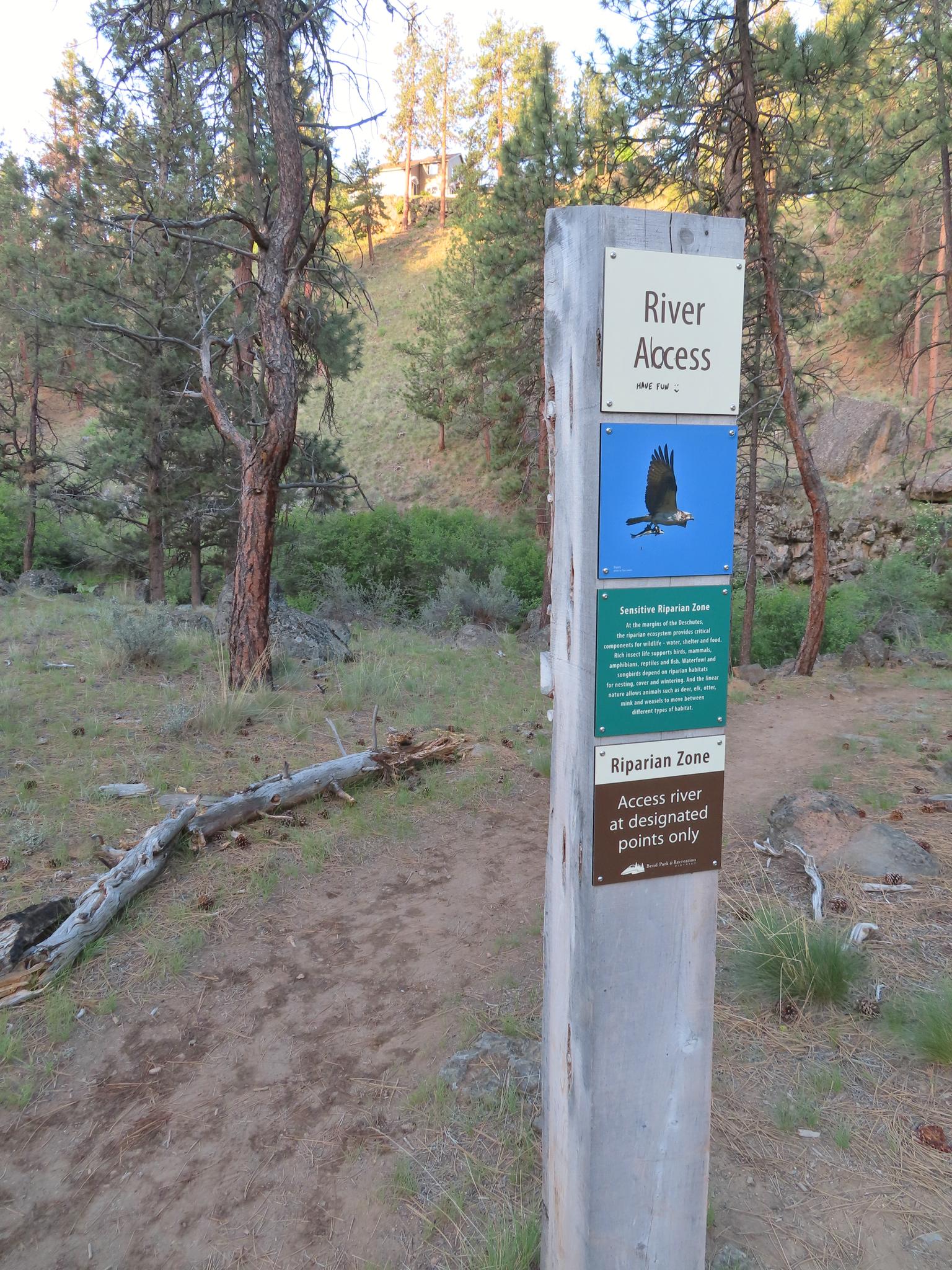 A sign post on a dirt path in a forest, indicating river access.