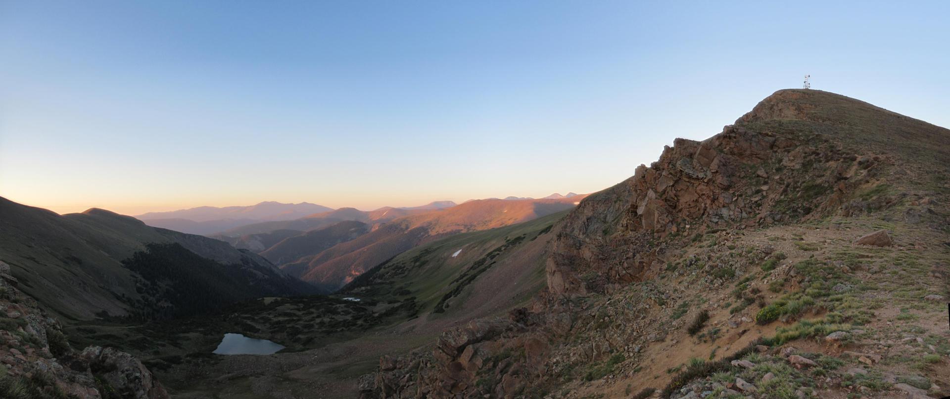 Mountain peak and valley bathed in warm sunrise light with a small lake.