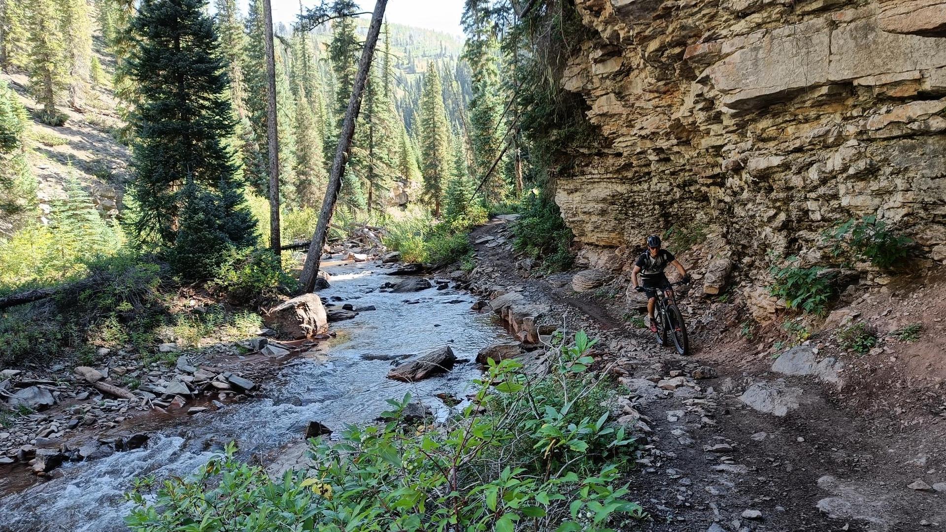 Mountain biker on a narrow trail beside a rocky stream and cliff face.