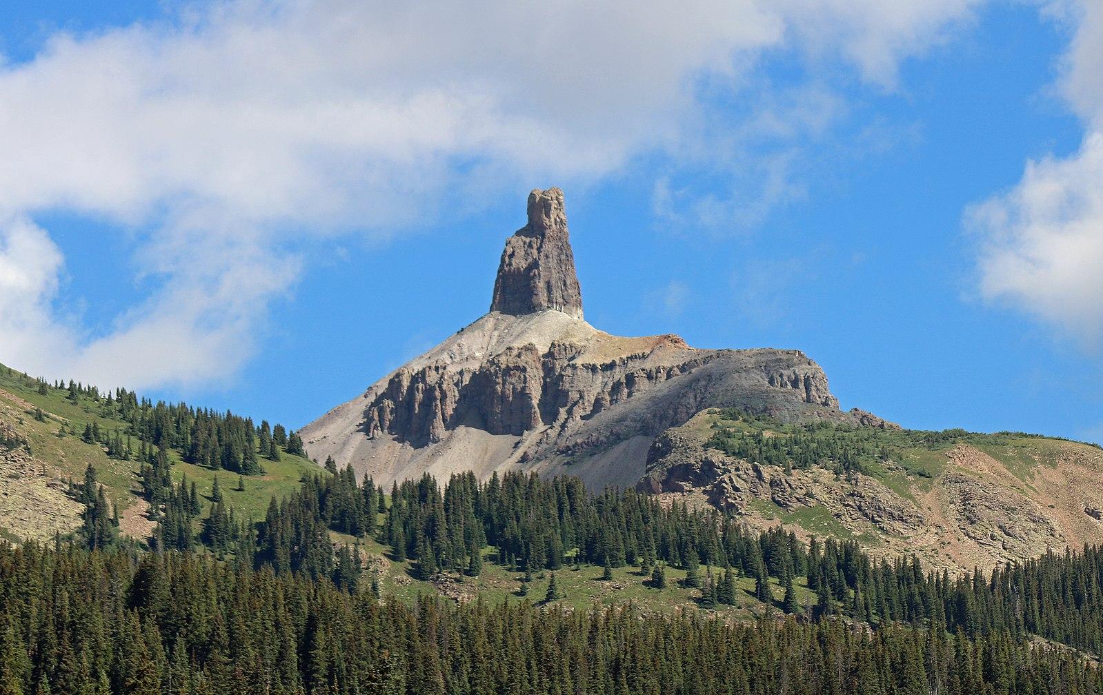 A unique, tall rock formation rises above a forested mountain under blue sky.