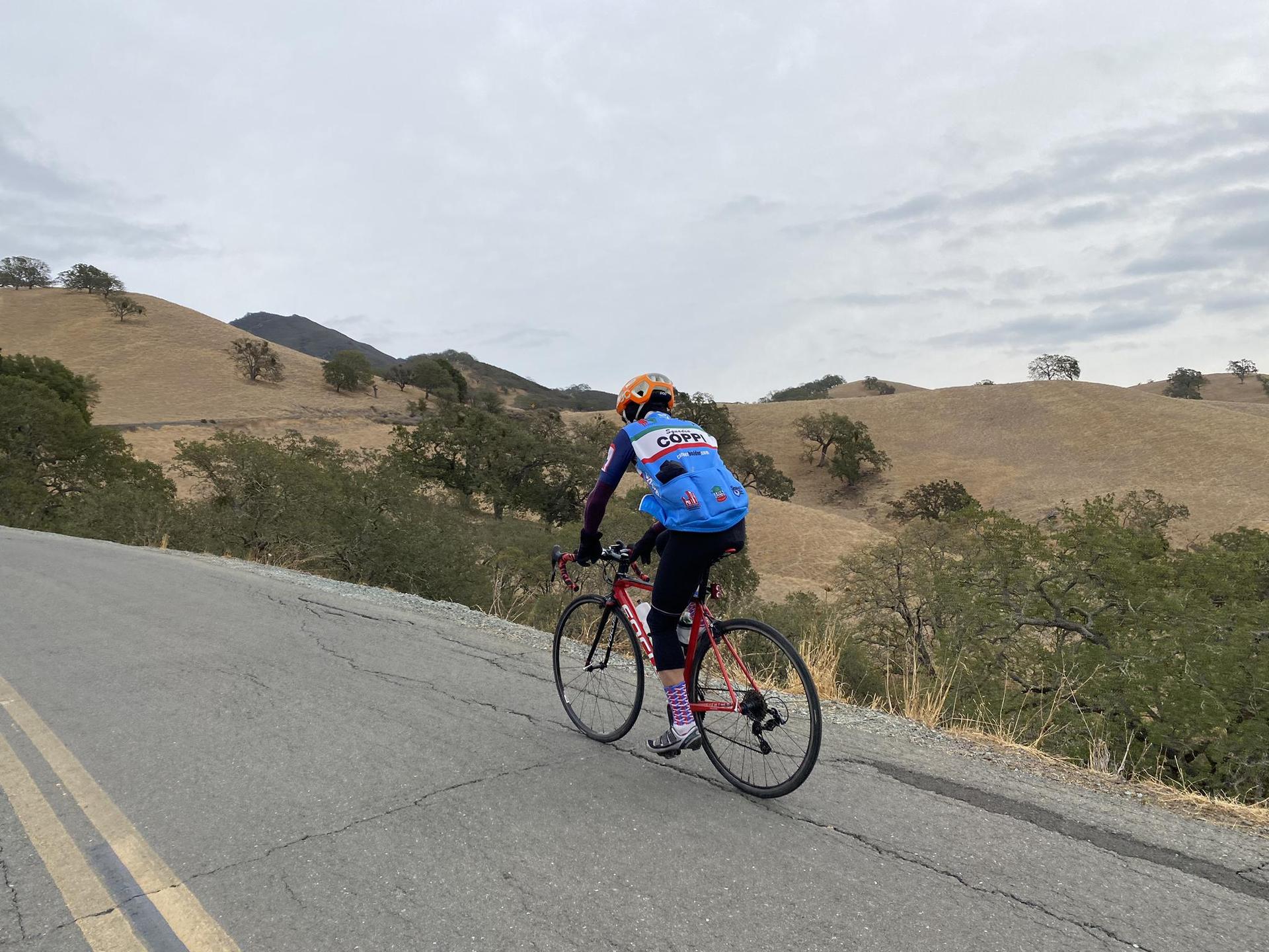 Cyclist in blue jersey rides uphill on a paved road with dry hills.