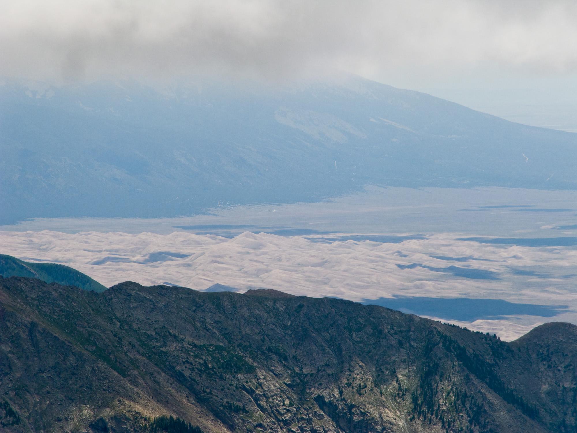 Distant sand dunes and mountains under a cloudy, hazy sky.
