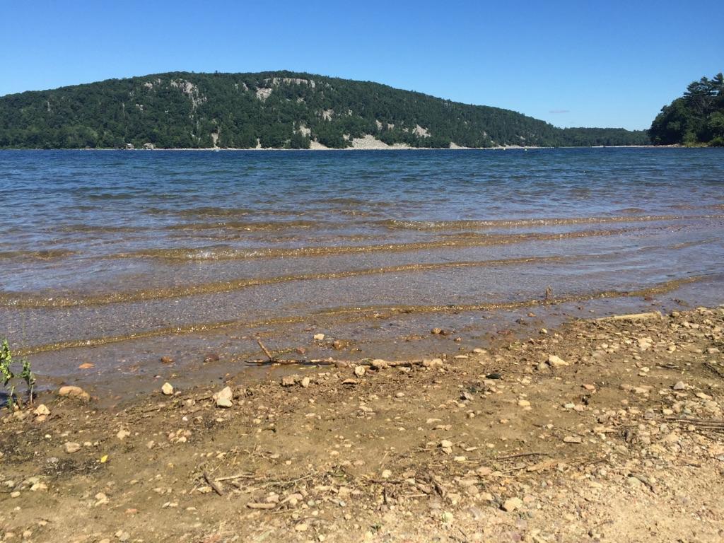 Clear blue lake with a rocky shore and green hills under bright sun.