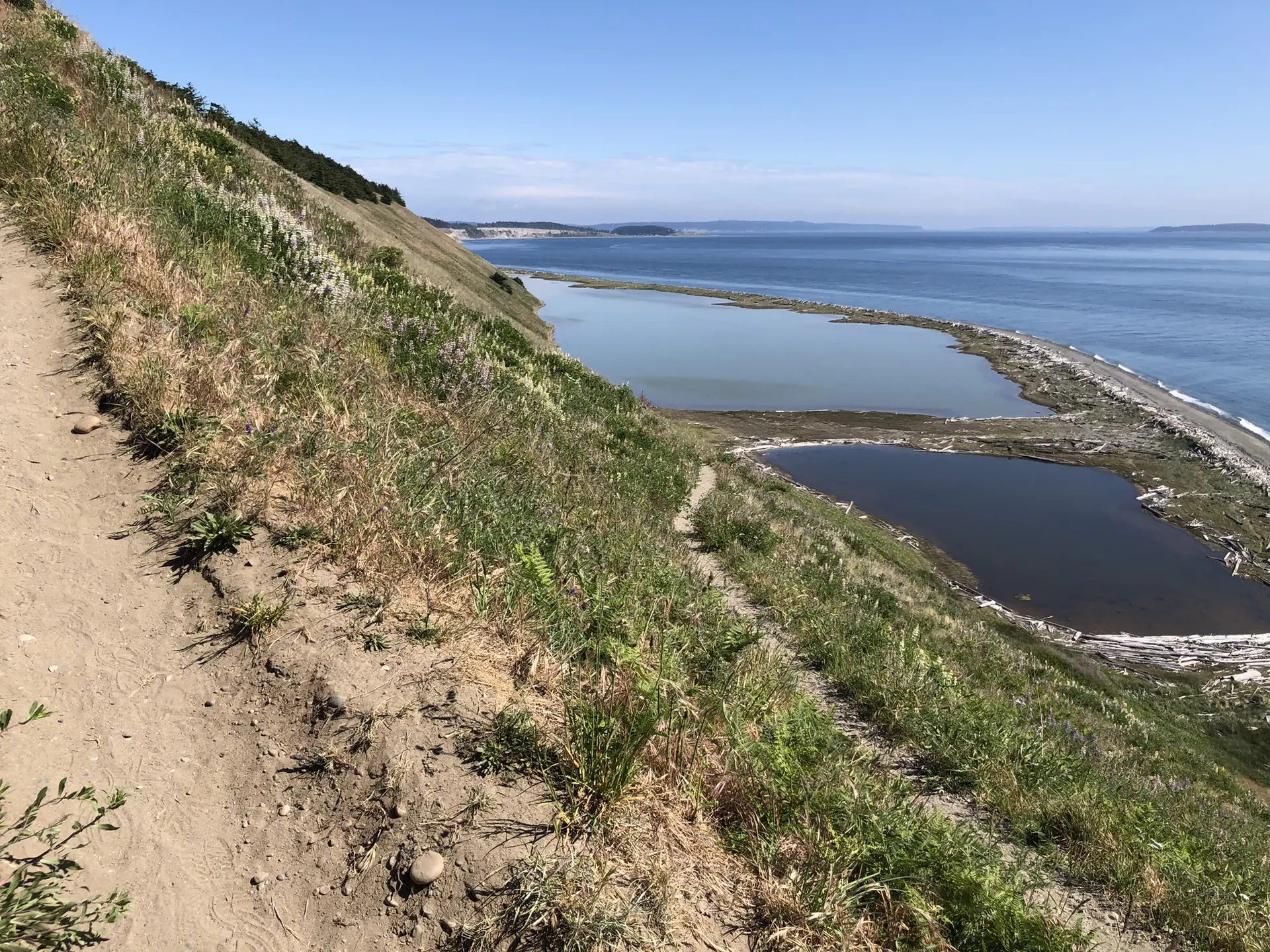 Dirt trail on a steep grassy slope overlooking two lagoons and the sea.