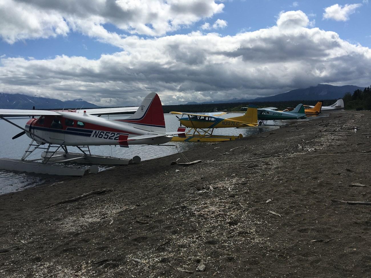 Several colorful seaplanes are parked on a dark gravel beach by a lake.