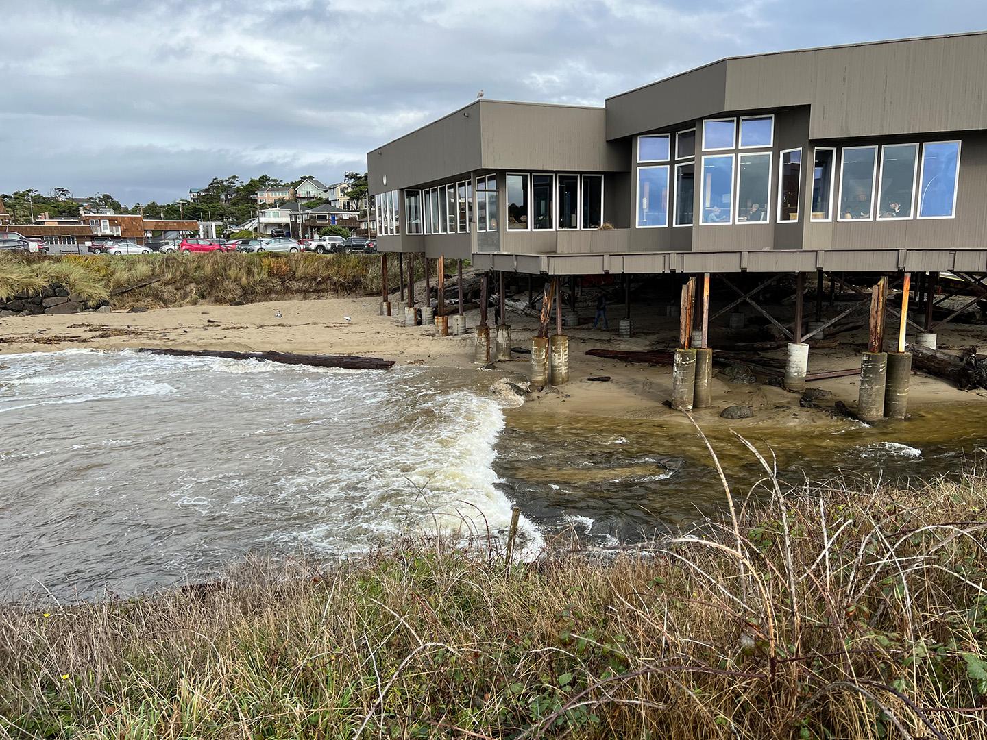 Modern building on stilts over water, with a sandy shore and overcast sky.