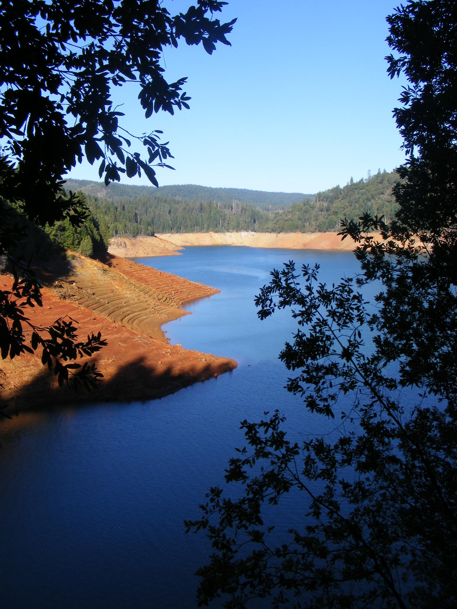 A blue lake with exposed reddish banks, framed by dark trees under a clear sky.