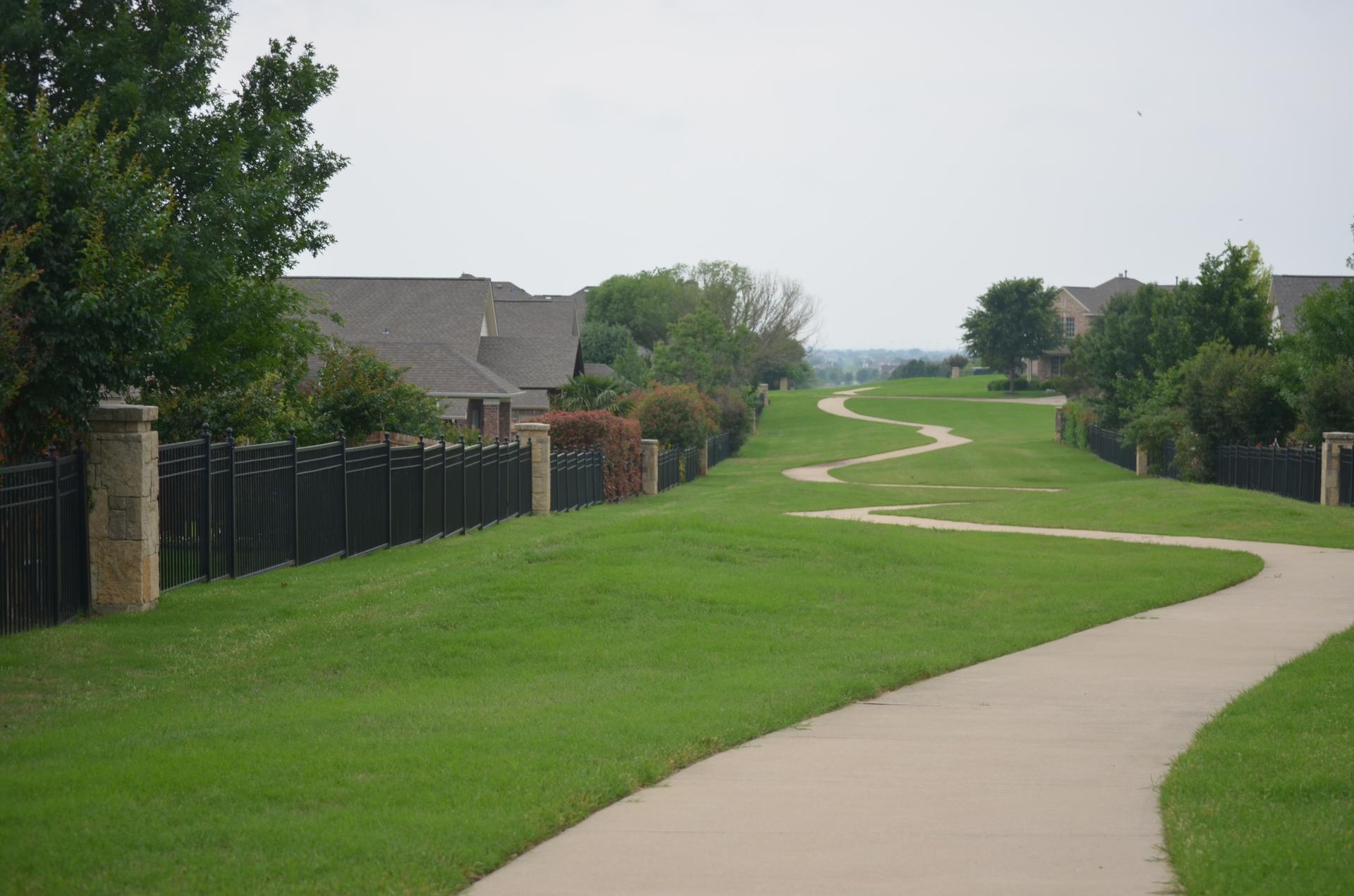 A winding paved path through green grass, bordered by a fence and houses.