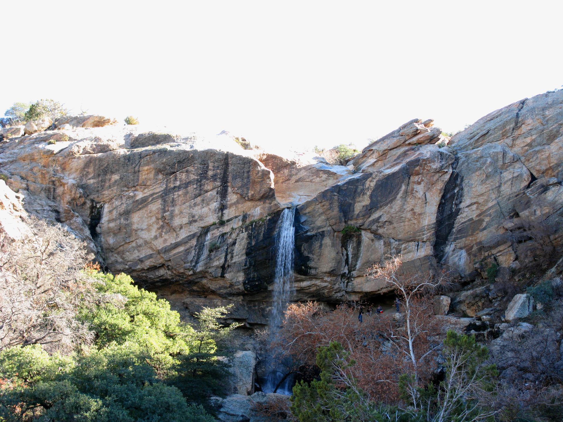 Bright sunlight illuminates a waterfall cascading down a tall, rocky canyon wall.