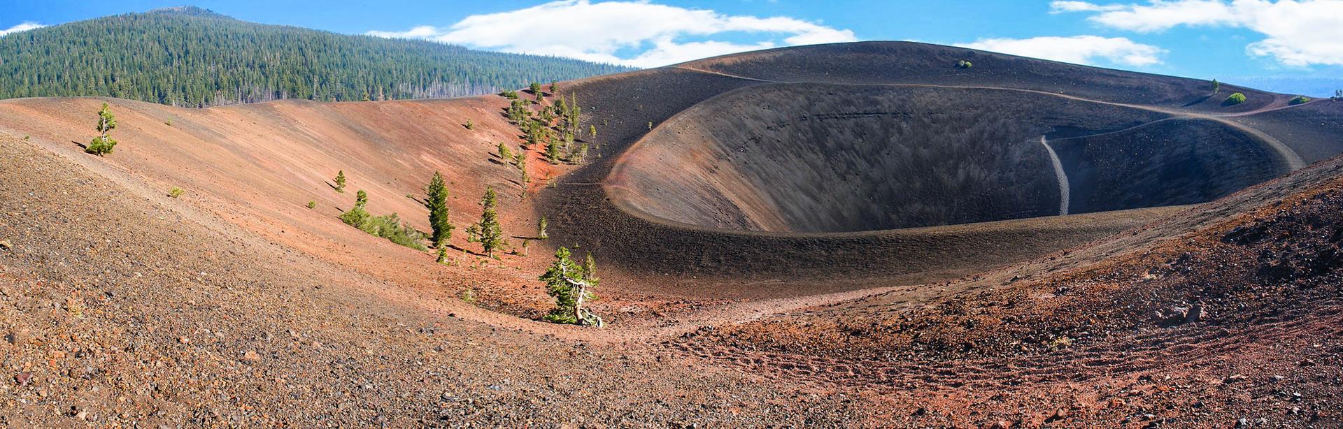 Panoramic view of a large, dark volcanic crater with reddish slopes and scattered trees.