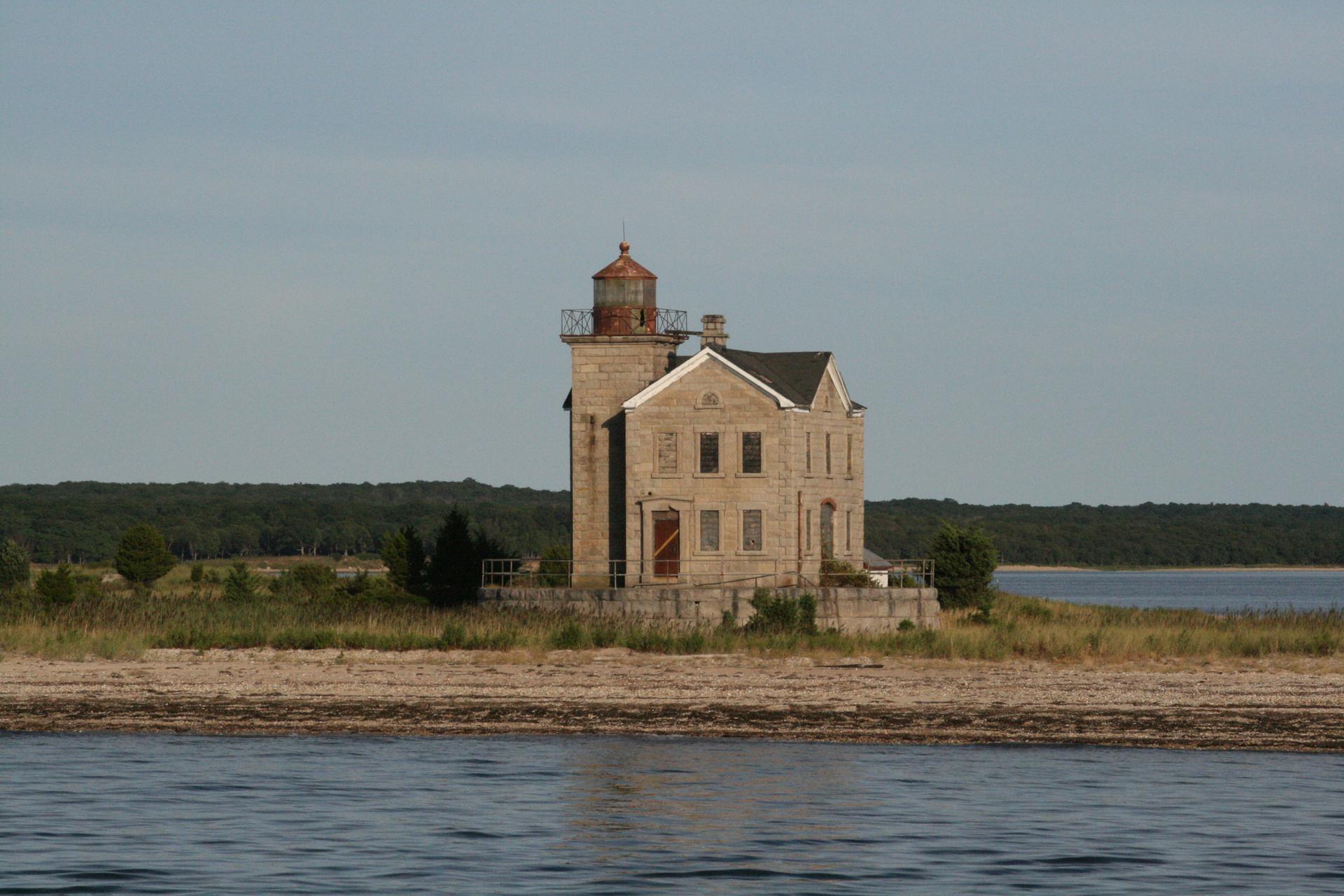 Old stone lighthouse on a sandy shore with water and forest under clear sky.