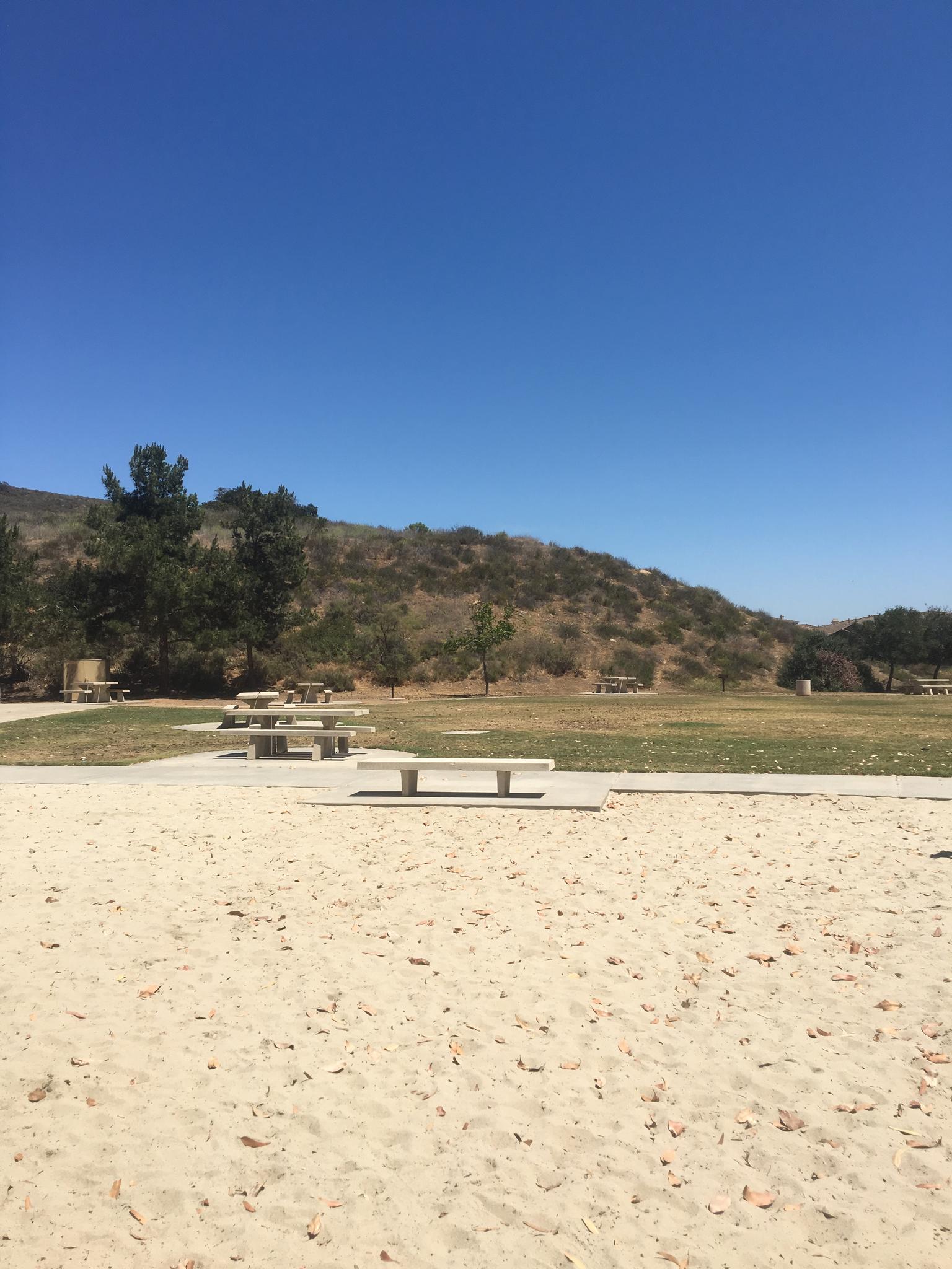 Sunny view of sandy ground, picnic tables, grassy field, and brushy hills.