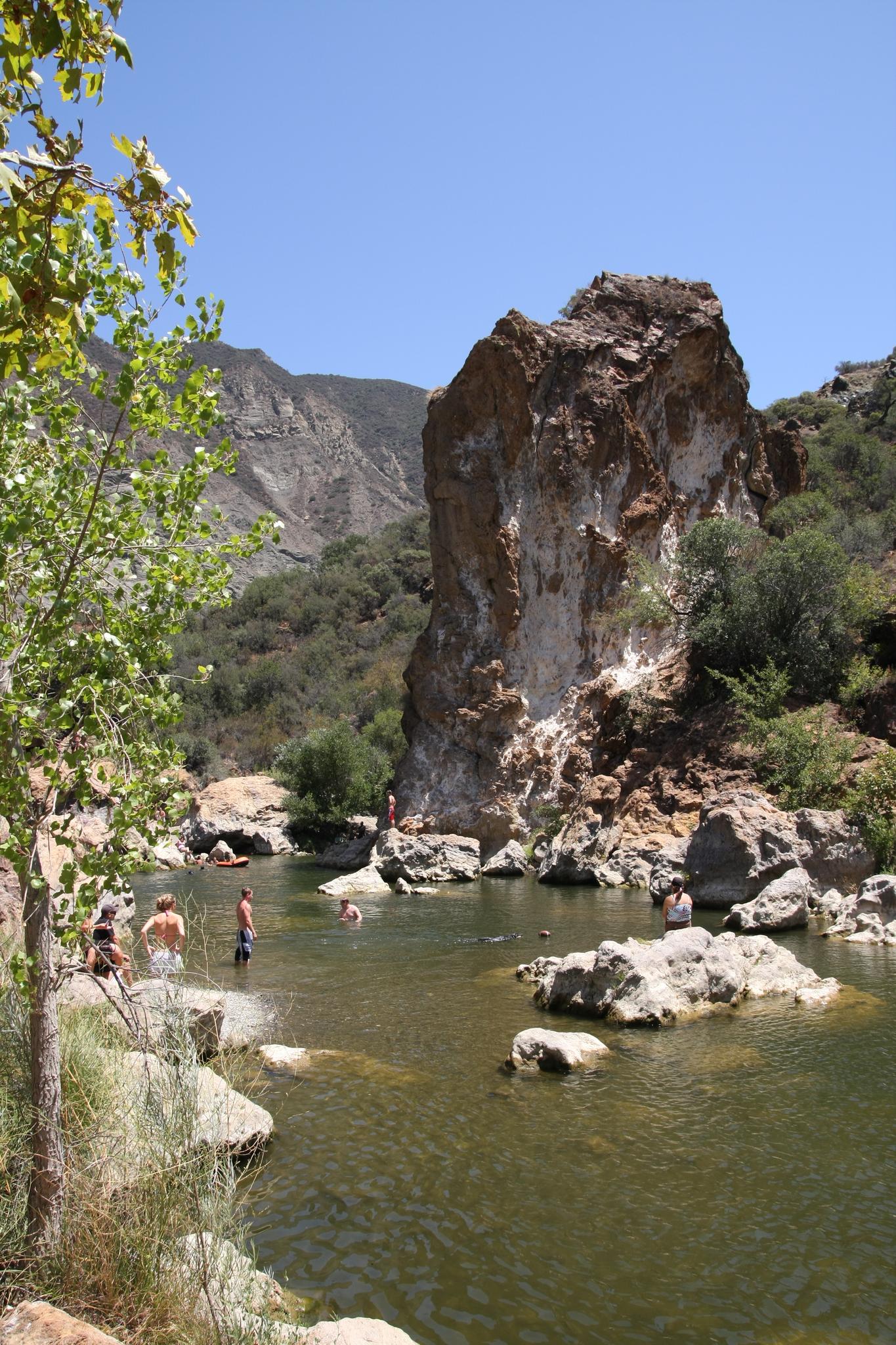 People swimming in a river next to a towering rock formation on a sunny day.