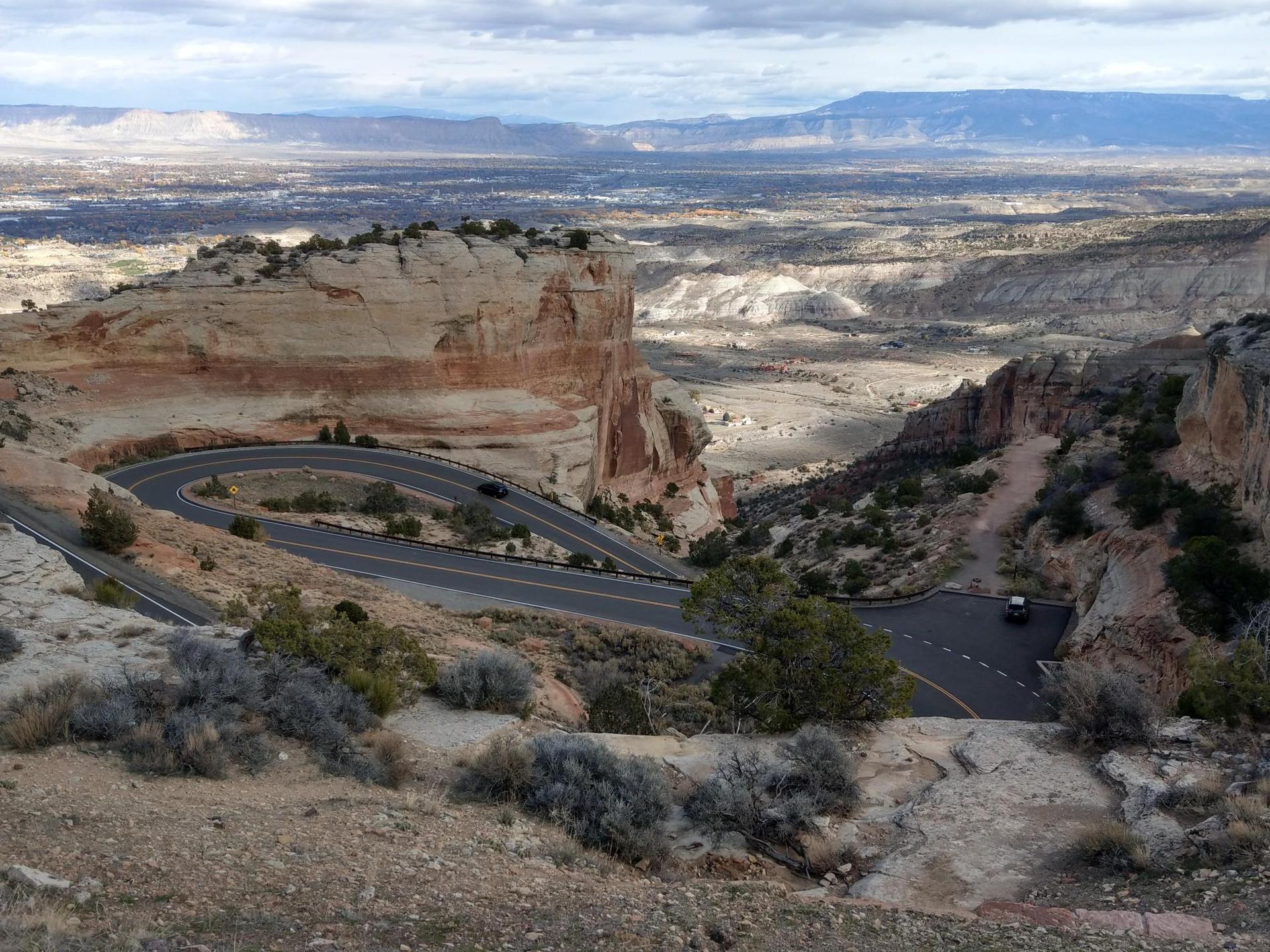 Winding road descends through a vast desert canyon under a cloudy sky.