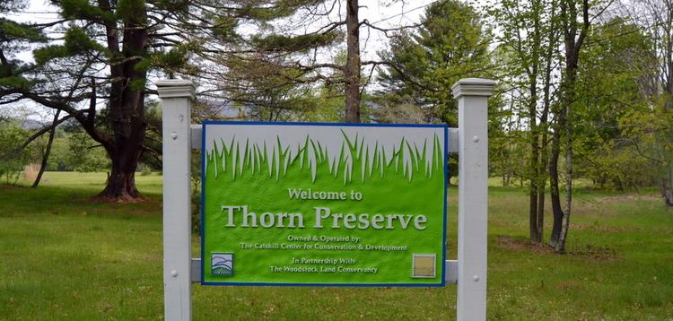 A green and white welcome sign stands in a grassy field with trees.