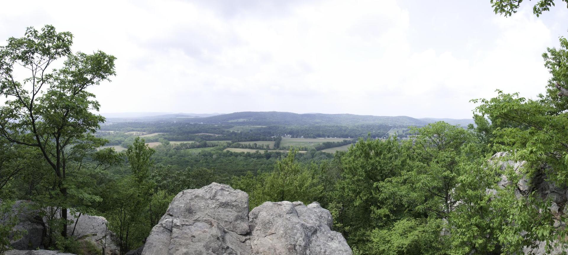 Panoramic view from rocky outcrop over green forests and distant hills under cloudy sky.