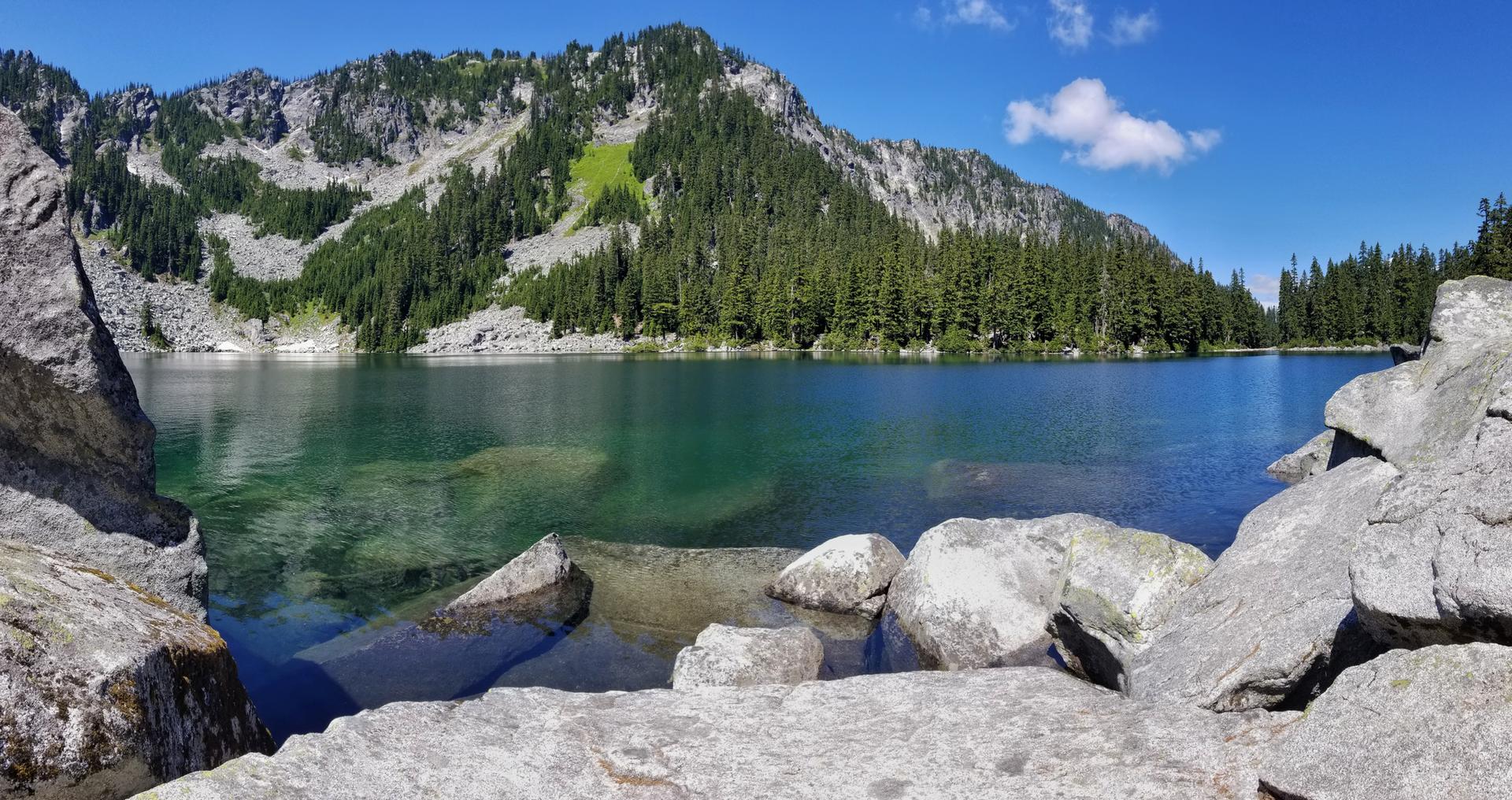 Clear mountain lake with rocky shores and forested peaks under a bright sky.