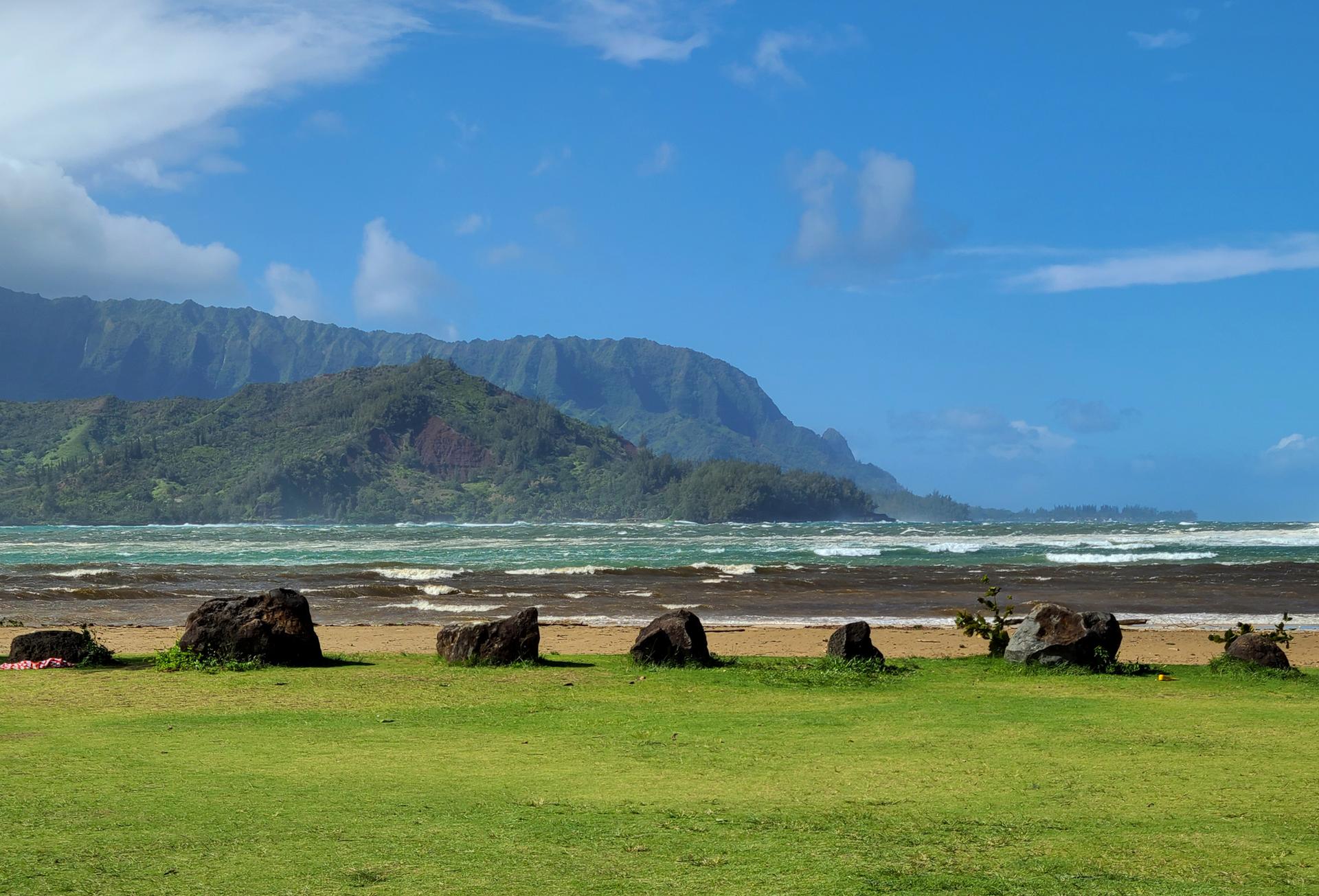 Grassy field meets a rocky beach with ocean and distant green mountains.