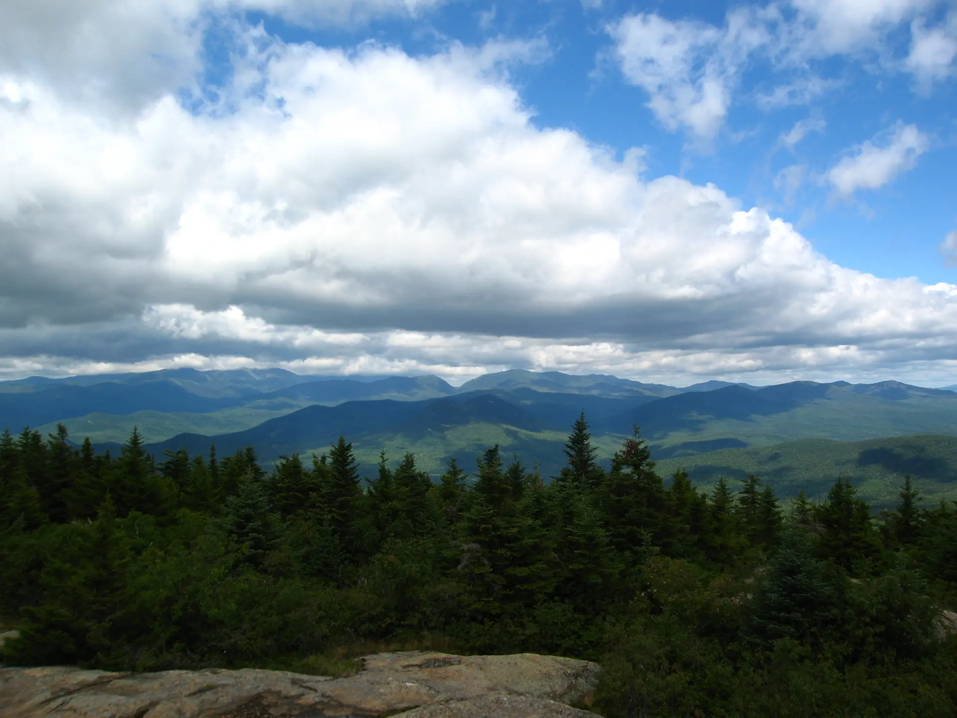 Green mountains and forests under a bright, cloudy sky from a rocky viewpoint.