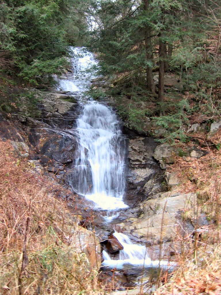A multi-tiered waterfall cascades down a rocky, forested slope under diffused light.