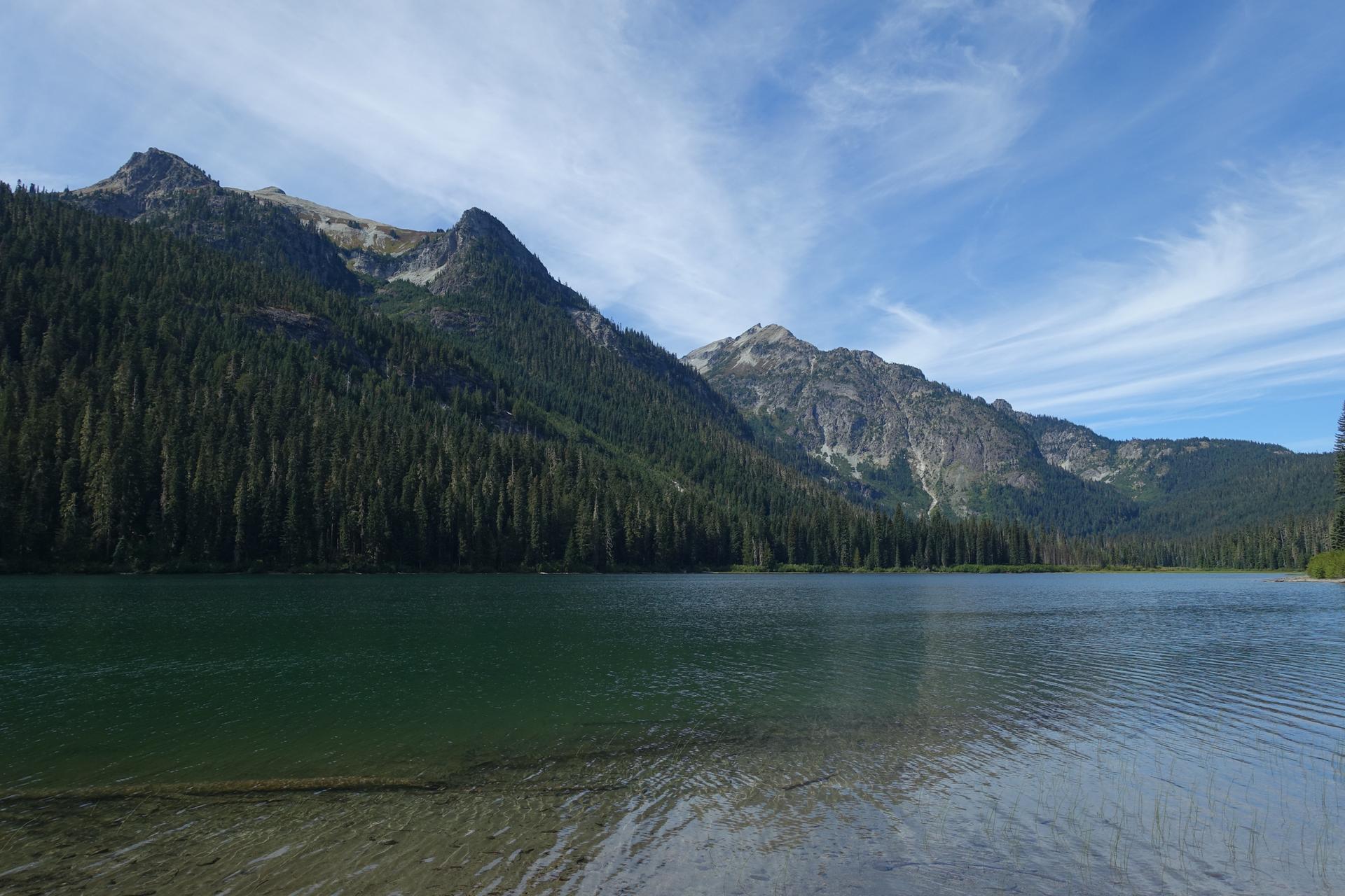 Calm lake reflecting dense evergreen forests and rocky mountains under a blue sky.