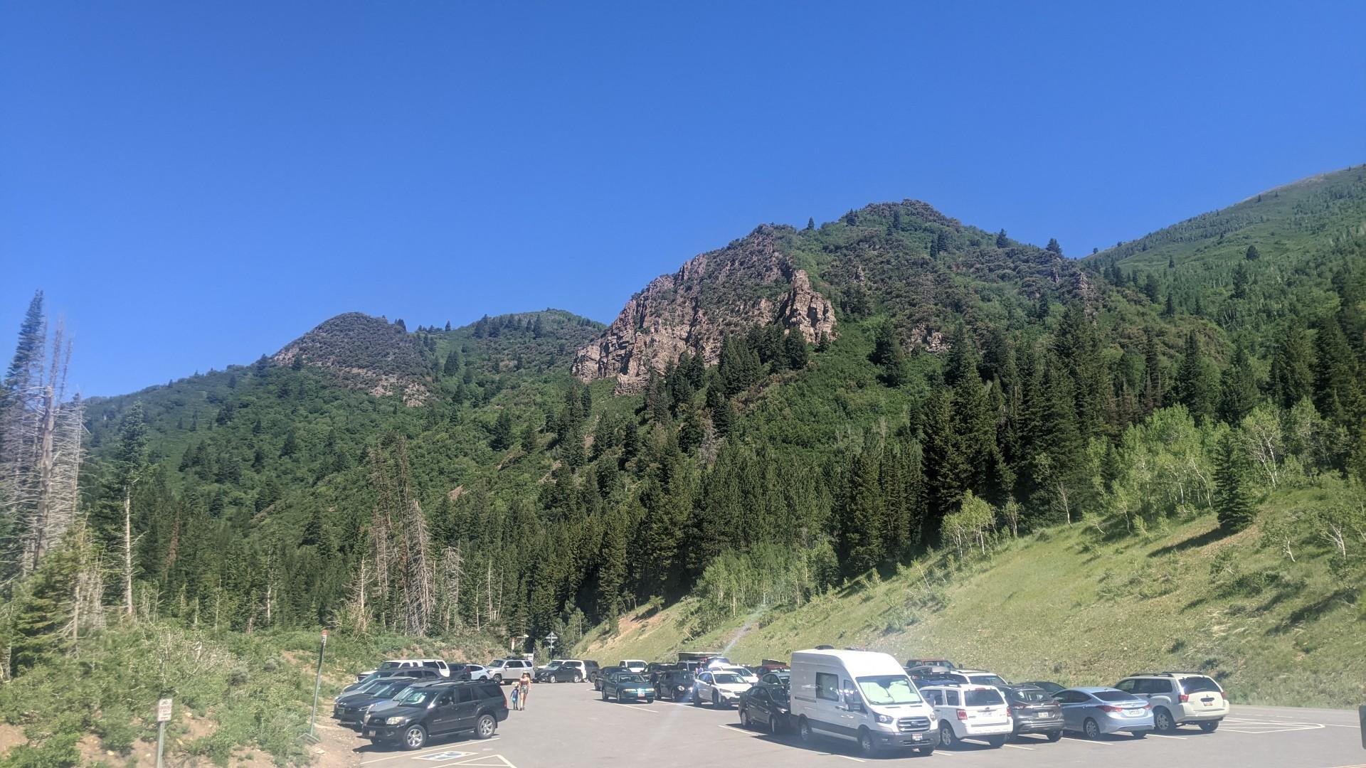 A parking lot full of vehicles at the base of sunny, forested mountains.