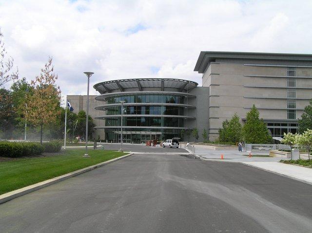 Modern building with circular glass entrance and paved driveway under a cloudy sky.