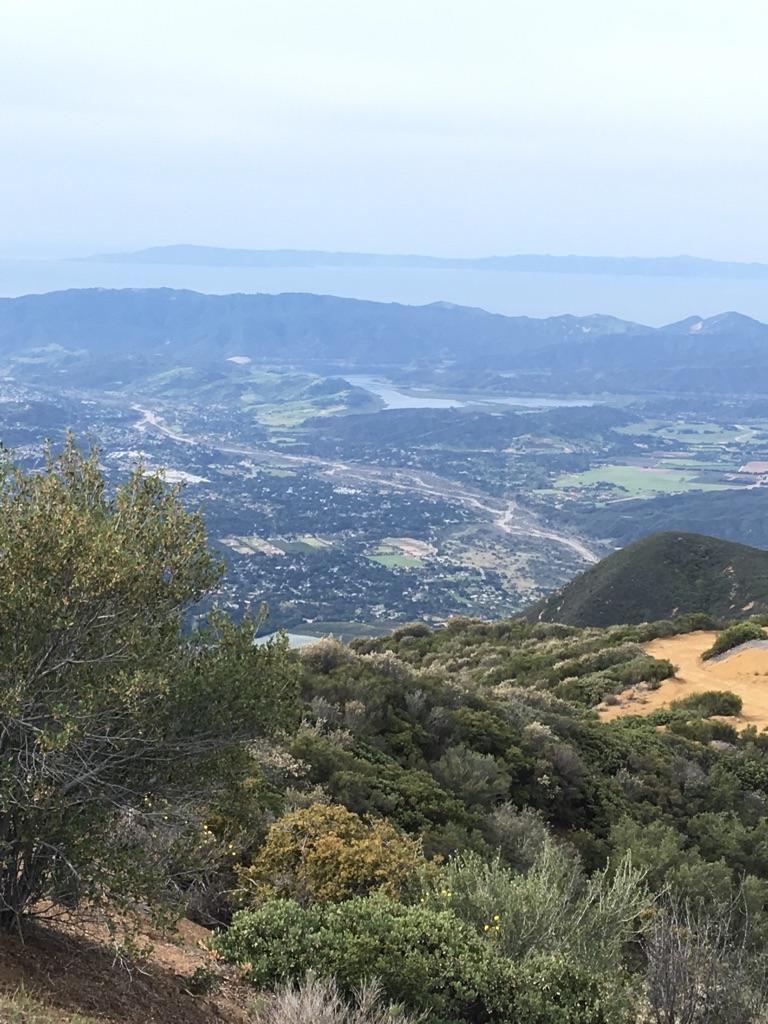 Expansive view of a valley, distant ocean, and mountains under hazy sky.